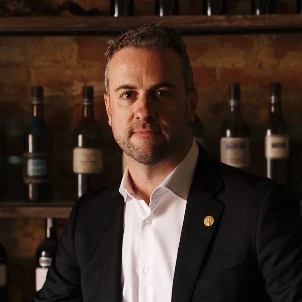 A man in a white shirt and dark blazer sitting in front of wine bottles on a wooden shelf.