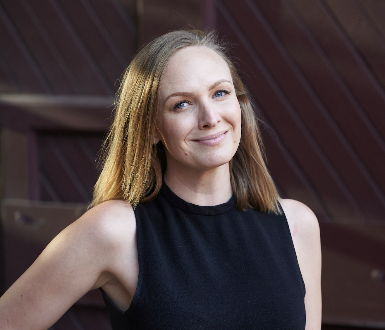 A woman with light brown hair, blue eyes, and light skin, smiling and wearing a black sleeveless top, standing outdoors in front of a wooden background.