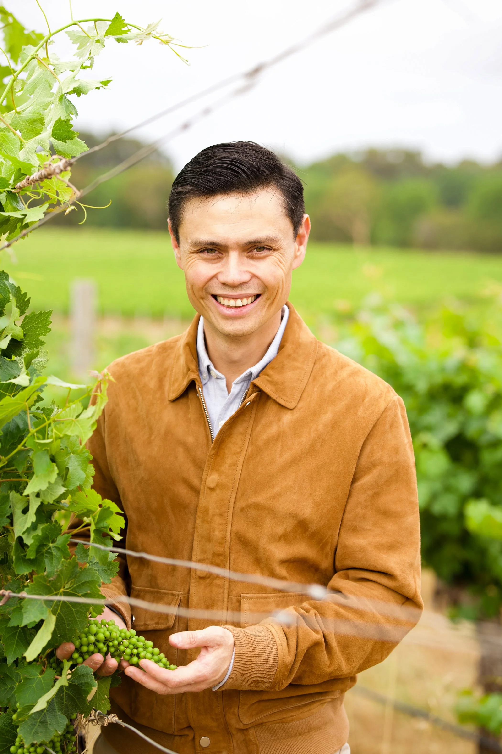 A man wearing a brown jacket and a gray shirt, standing in a vineyard, holding a cluster of green grapes.