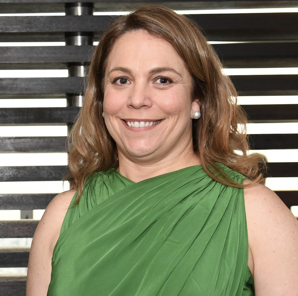 A woman with light skin, shoulder-length auburn hair, wearing a green dress with a pleated design, pearl earrings, smiling in front of black horizontal blinds.