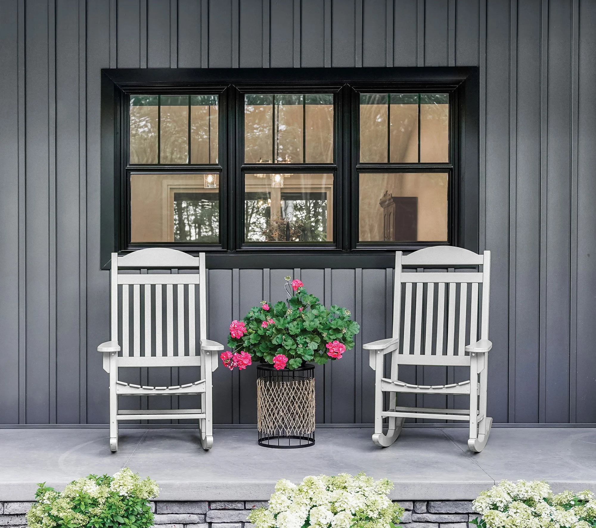 Two white rocking chairs with a pink flowering plant between them on a porch in front of a dark gray wall with black-framed window.