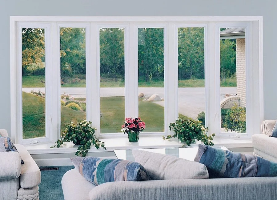 Living room with large window overlooking a yard with green grass, trees, and a brick house, decorated with potted plants and a sofa with blue and beige cushions.