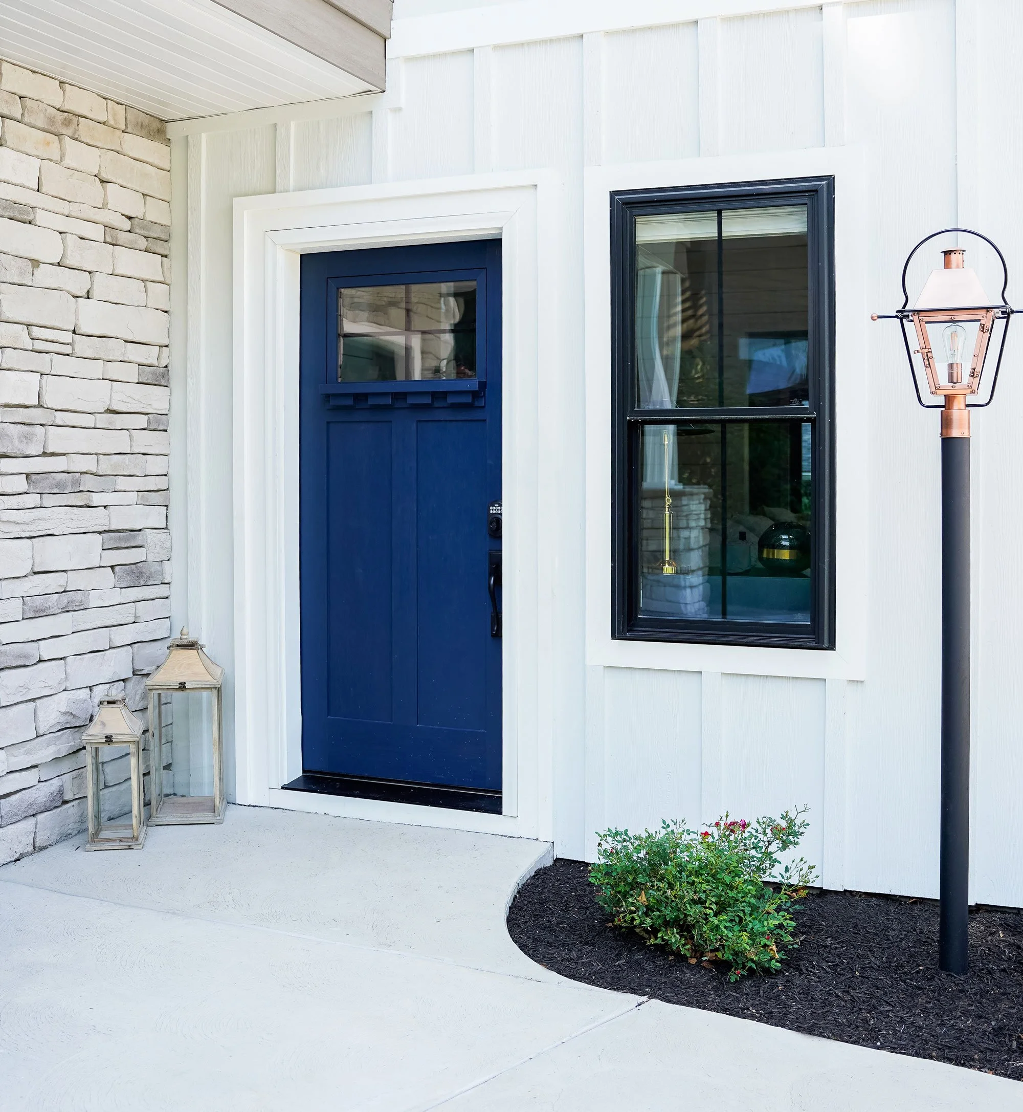 Front porch entrance with a blue door, black-framed window, lantern-style lamp post, and two lantern decorations on the ground, surrounded by a small garden bed.