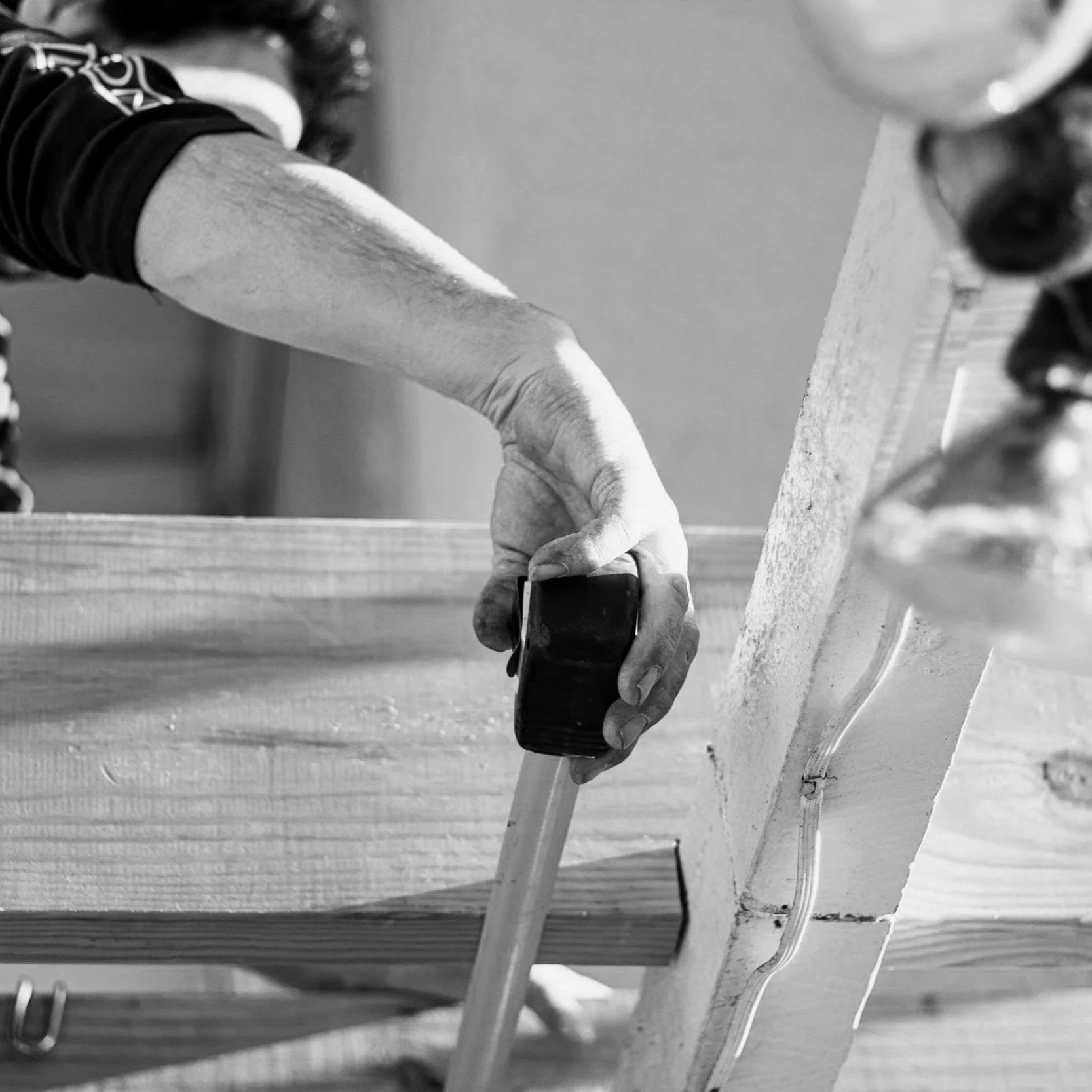 A person's hand holding a tape measure measuring a piece of wood placed on a workbench in a woodworking workshop.