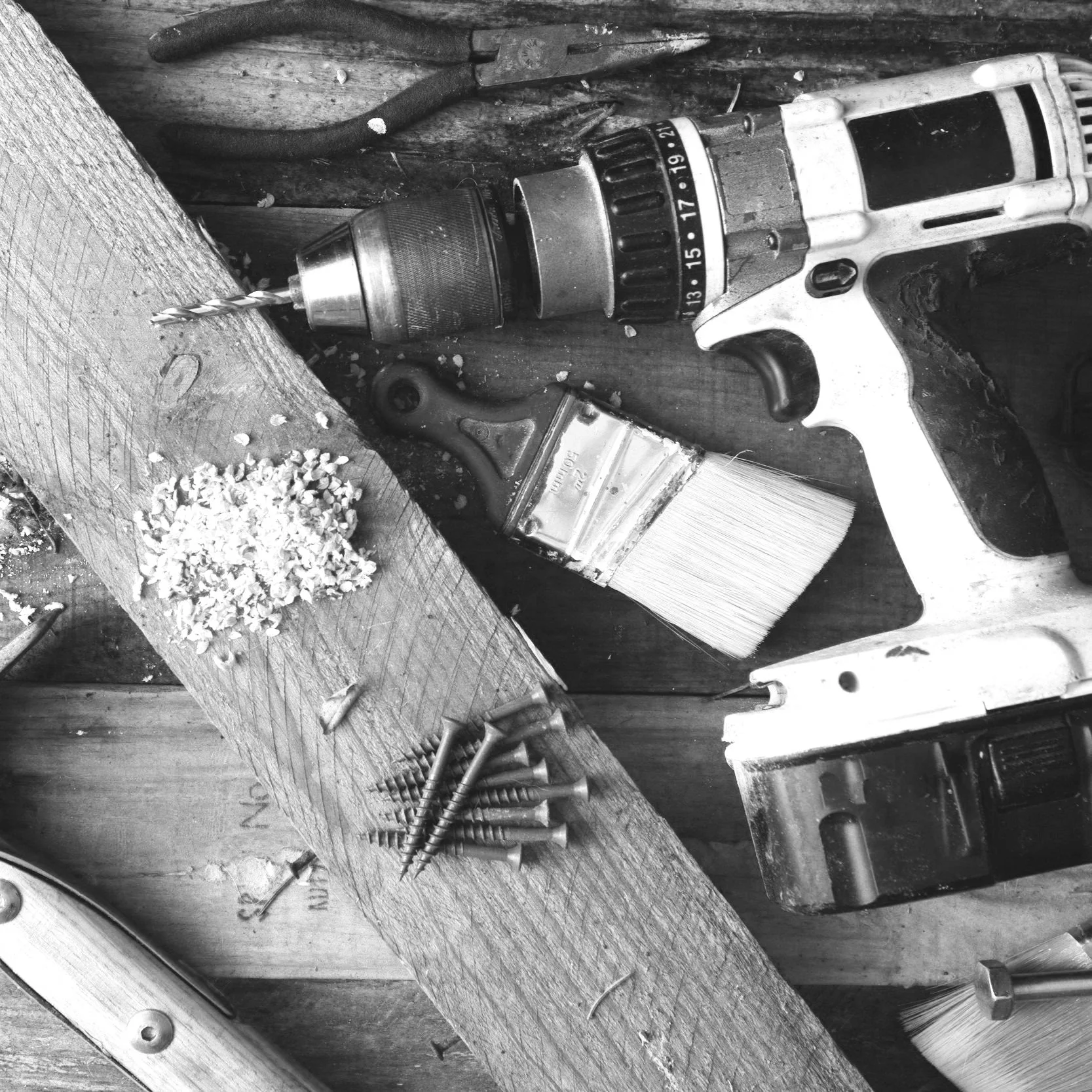 A black and white photo of a power drill, a paintbrush, nails and screws, wood, and other tools on a wooden surface in a woodworking setting.