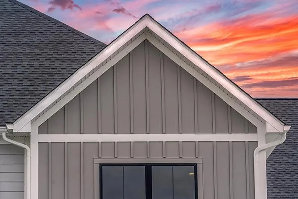 Close-up of the front gable of a house with gray vertical siding and a black window, under a colorful sunset sky.