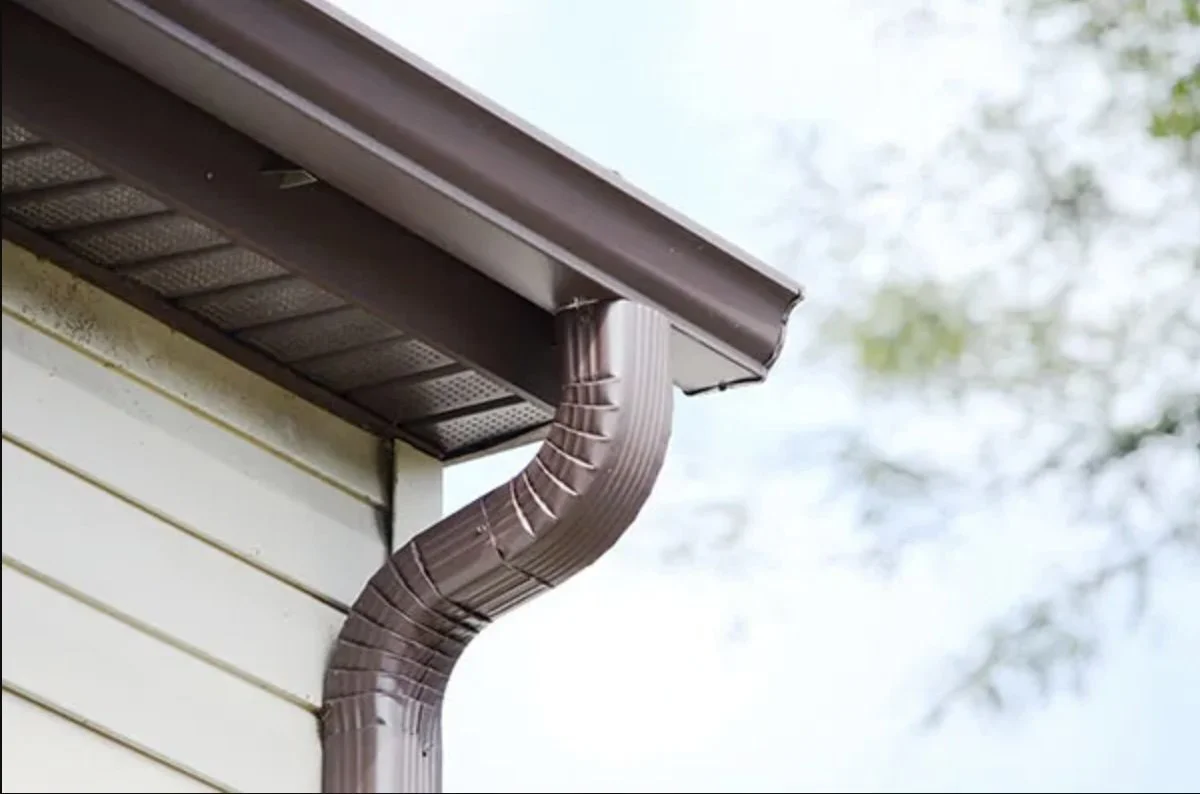 Close-up view of a house's brown metal gutter and downspout attached to a beige siding, with a blurred background of sky and trees.