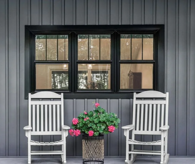 Two white wooden chairs with armrests and slatted backs on a porch, flanking a potted pink geranium plant, in front of a black paneled wall with a window showing reflections of trees and sky.