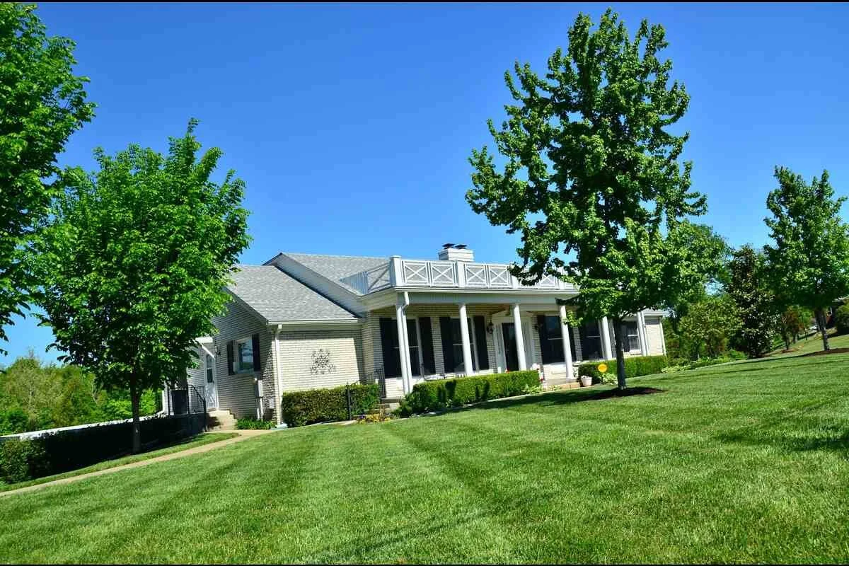 A large two-story house with a front porch, surrounded by green trees and well-maintained lawn, under a clear blue sky.