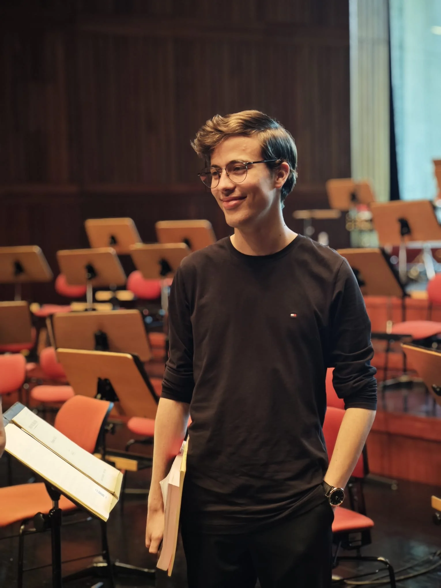 A young man with glasses and a watch standing in an orchestra hall, holding sheet music and smiling.