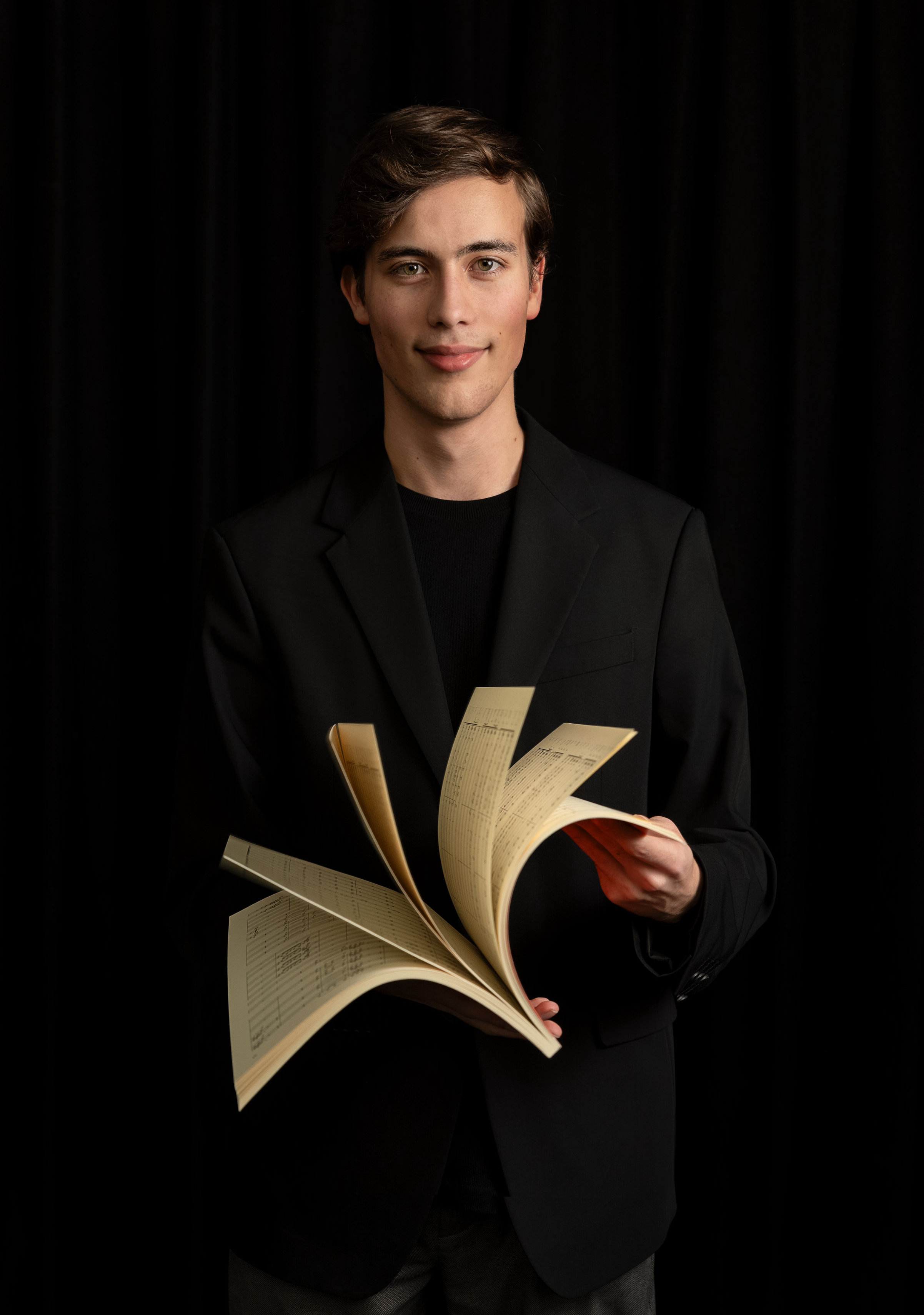 Young man with brown hair wearing a black blazer and black shirt holding an open book against a dark background.