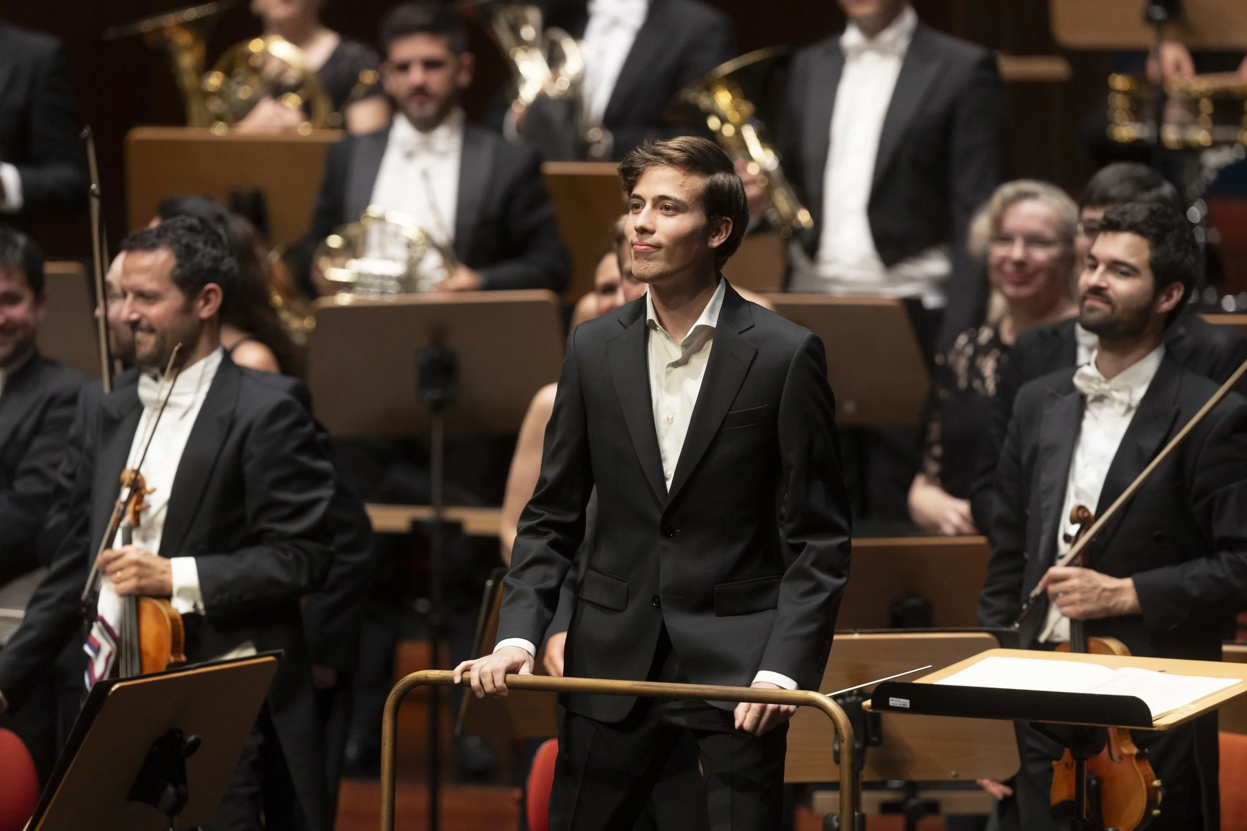 Conductor in black tuxedo leading an orchestra during a performance, with orchestra members holding string and wind instruments behind him.