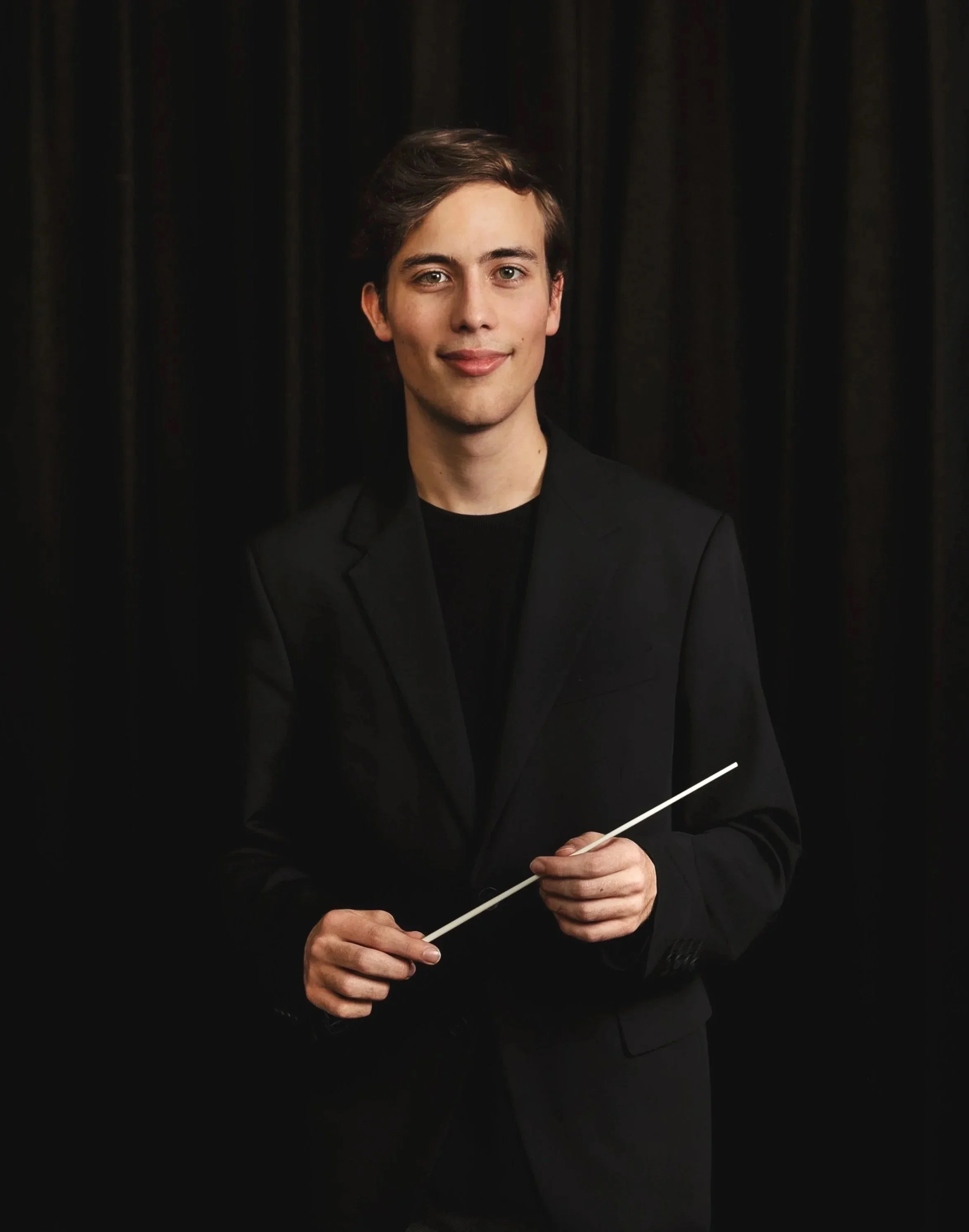 Young male orchestra conductor holding a baton, wearing a black suit, standing in front of a dark curtain.