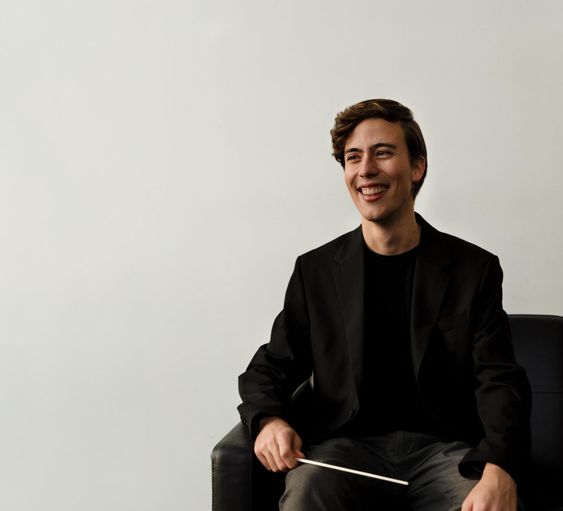 A young man sitting on a black chair, smiling and looking to his right, holding a baton in his right hand, wearing a black blazer and gray pants, against a plain white wall background.