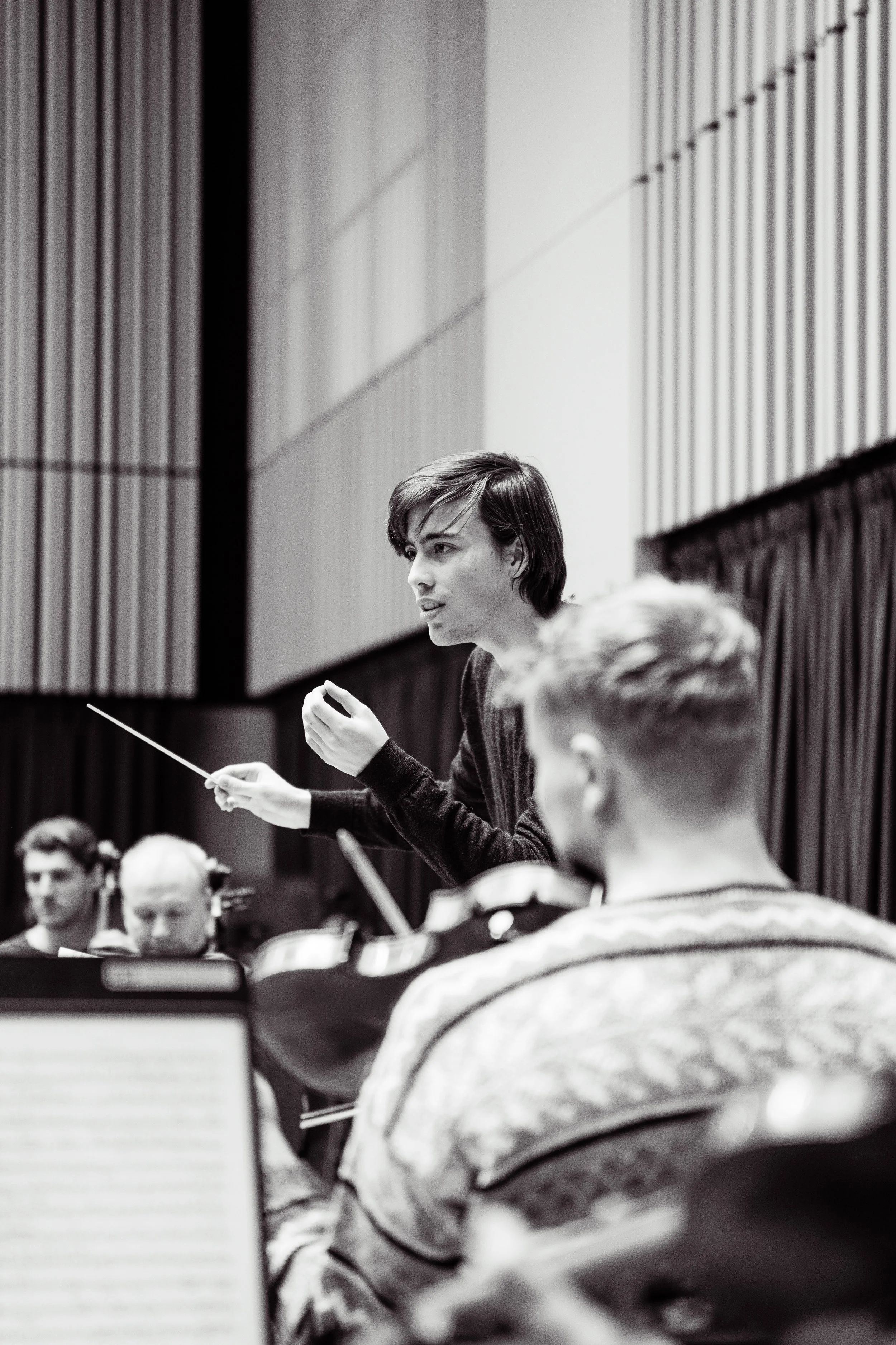 Conductor leading an orchestra during rehearsal, with musicians and sheet music in front of them, in a concert hall.