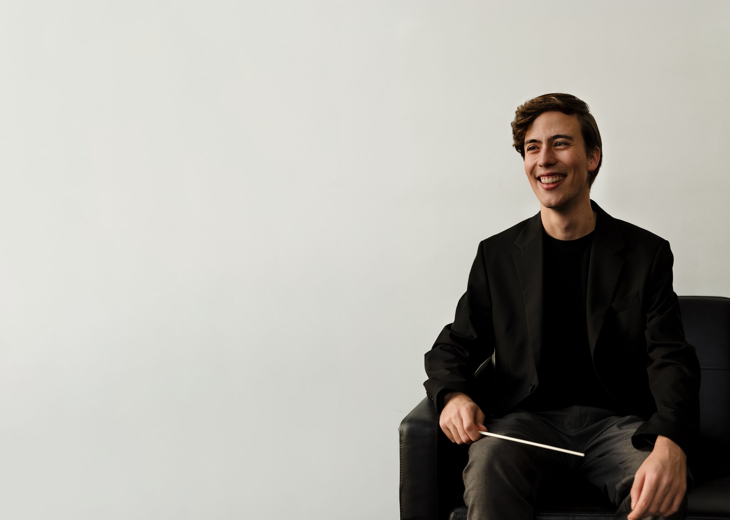 A young man with brown hair, wearing a black blazer and black shirt, sitting on a black chair, smiling, holding a conductor's baton in his right hand, in front of a plain white wall.