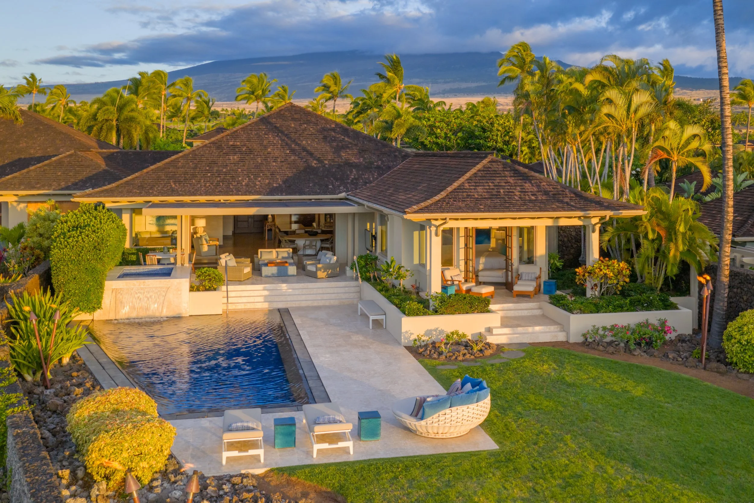Luxury house with outdoor patio, pool, and lush greenery, surrounded by numerous palm trees, with a mountain and cloudy sky in the background.