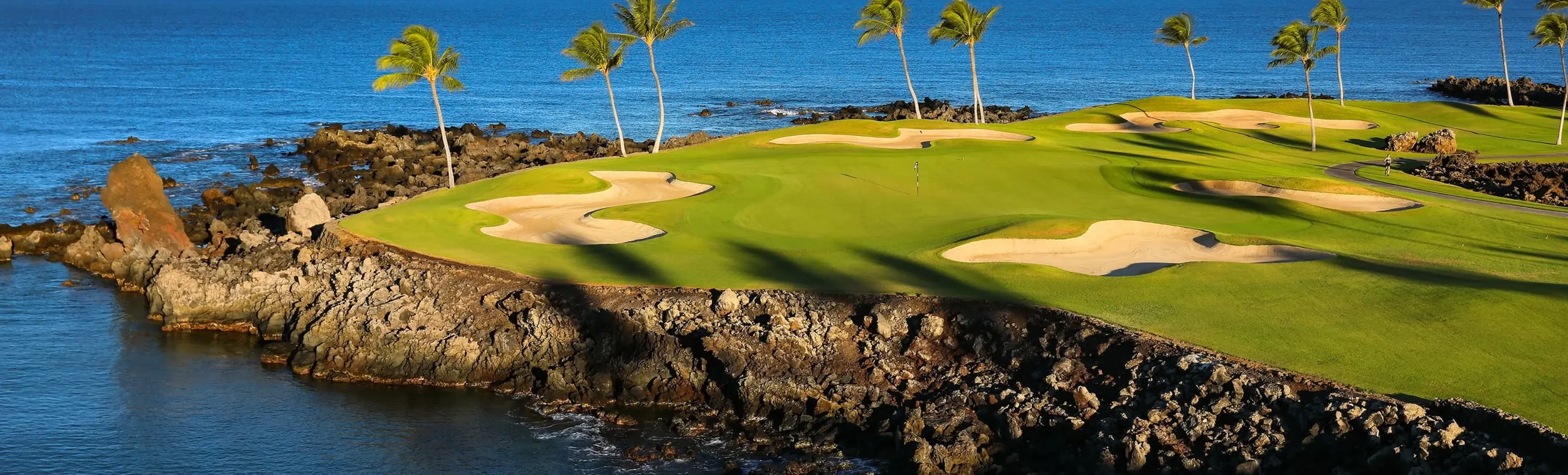 A coastal golf course with well-maintained green fairways, sand bunkers, and palm trees overlooking the ocean with rocky shoreline.