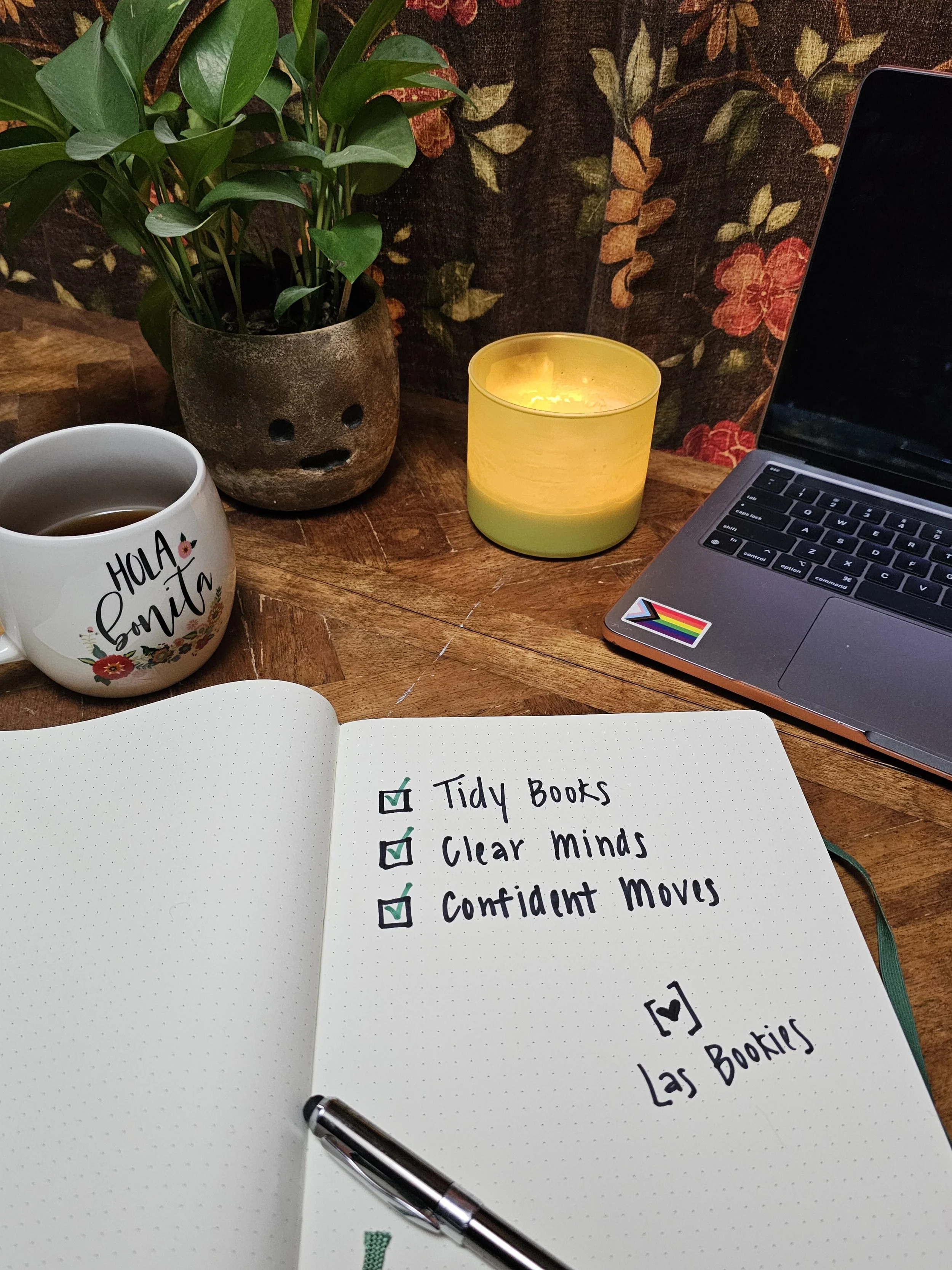 A workspace with a potted green plant, a lit yellow candle, a gray laptop with a rainbow pride sticker, a white mug with the words 'Hola Sonja' filled with coffee, and an open dotted notebook with a checklist that reads 'Tidy Books', 'Clear Minds', 'Confident Moves', signed 'Las Bookies'.