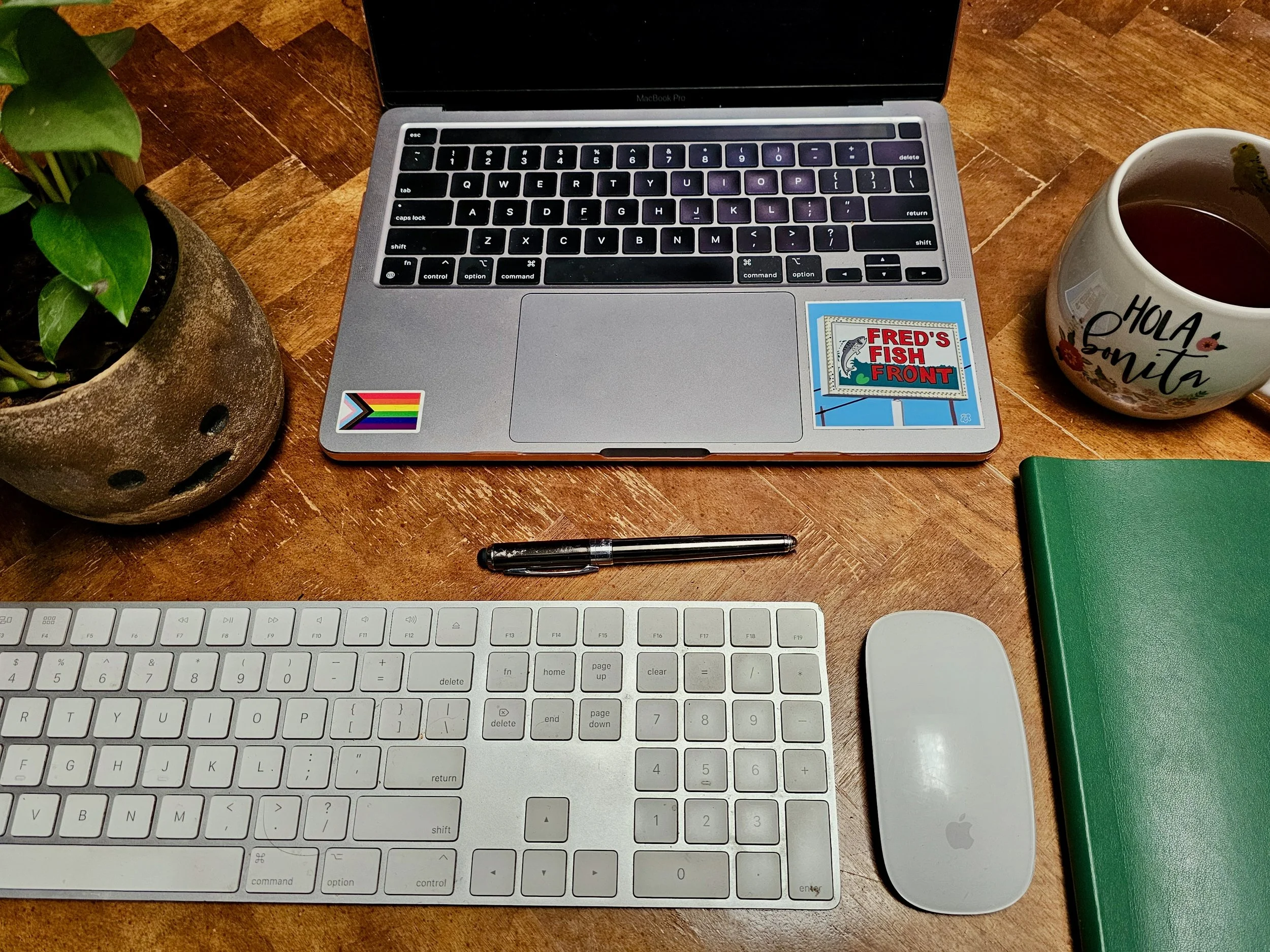 A workspace featuring a MacBook Pro laptop, a wireless Apple keyboard, a wireless Apple mouse, a black pen, a green notebook, a potted plant with a smiling face, a white mug with