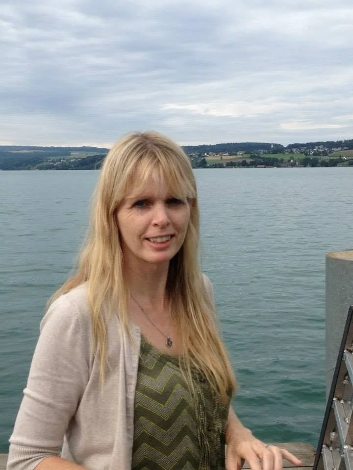 A woman with long blonde hair standing near a body of water with hills in the background, wearing a light-colored cardigan over a patterned green top.