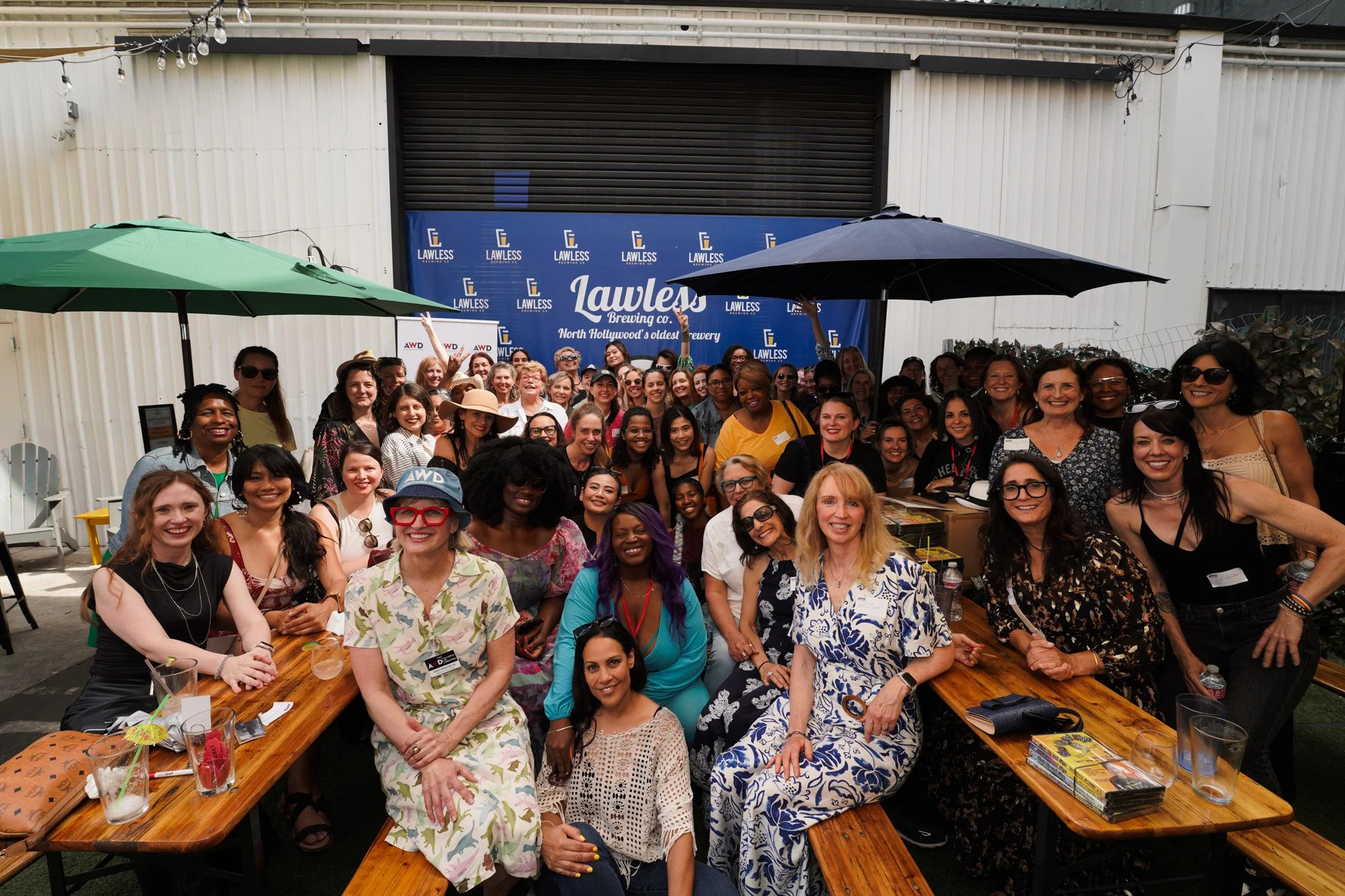 Group of women gathered at an outdoor event with a backdrop that reads 'Lawless Brewing Co.', some seated and some standing, with umbrellas providing shade.