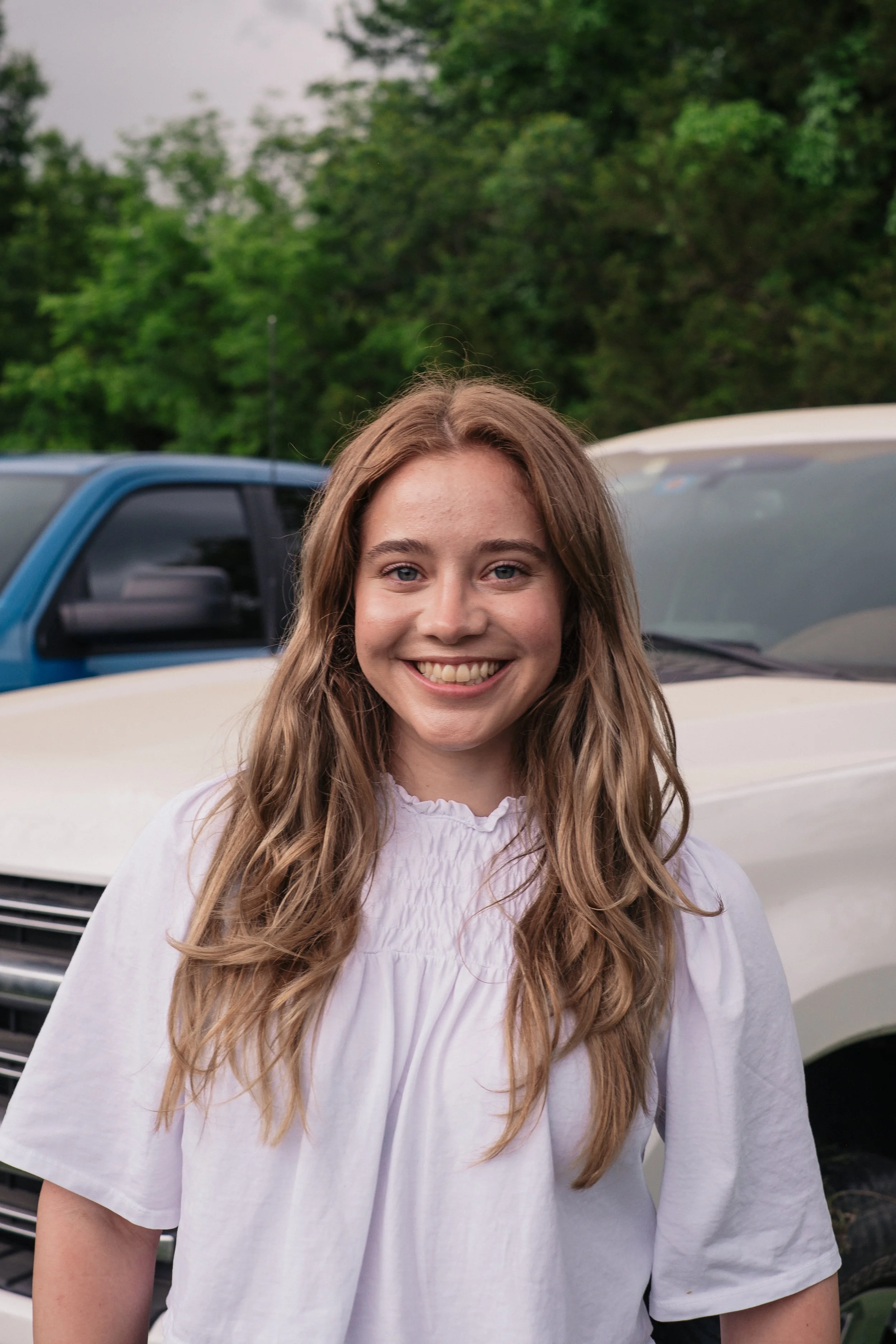 A young woman with long, curly light brown hair smiling in front of parked cars and green trees.