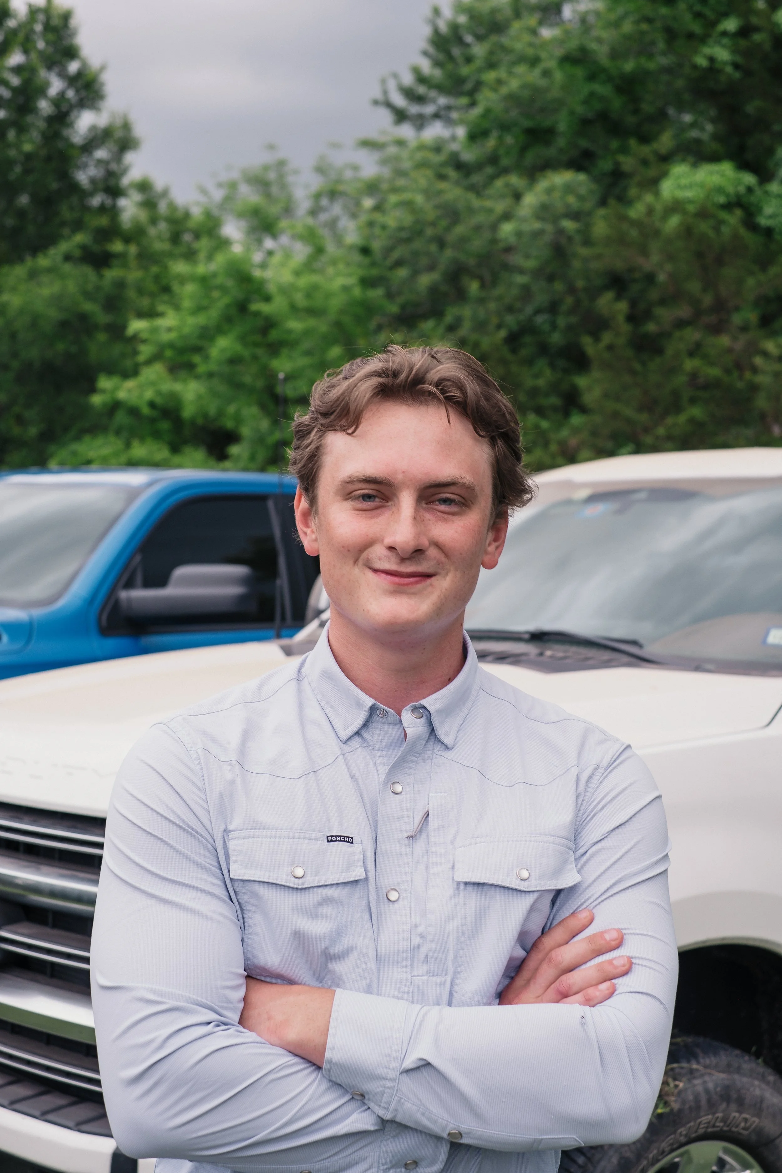 A young man with light brown hair, wearing a light blue button-up shirt with crossed arms, stands in front of a white vehicle with another vehicle and trees in the background.