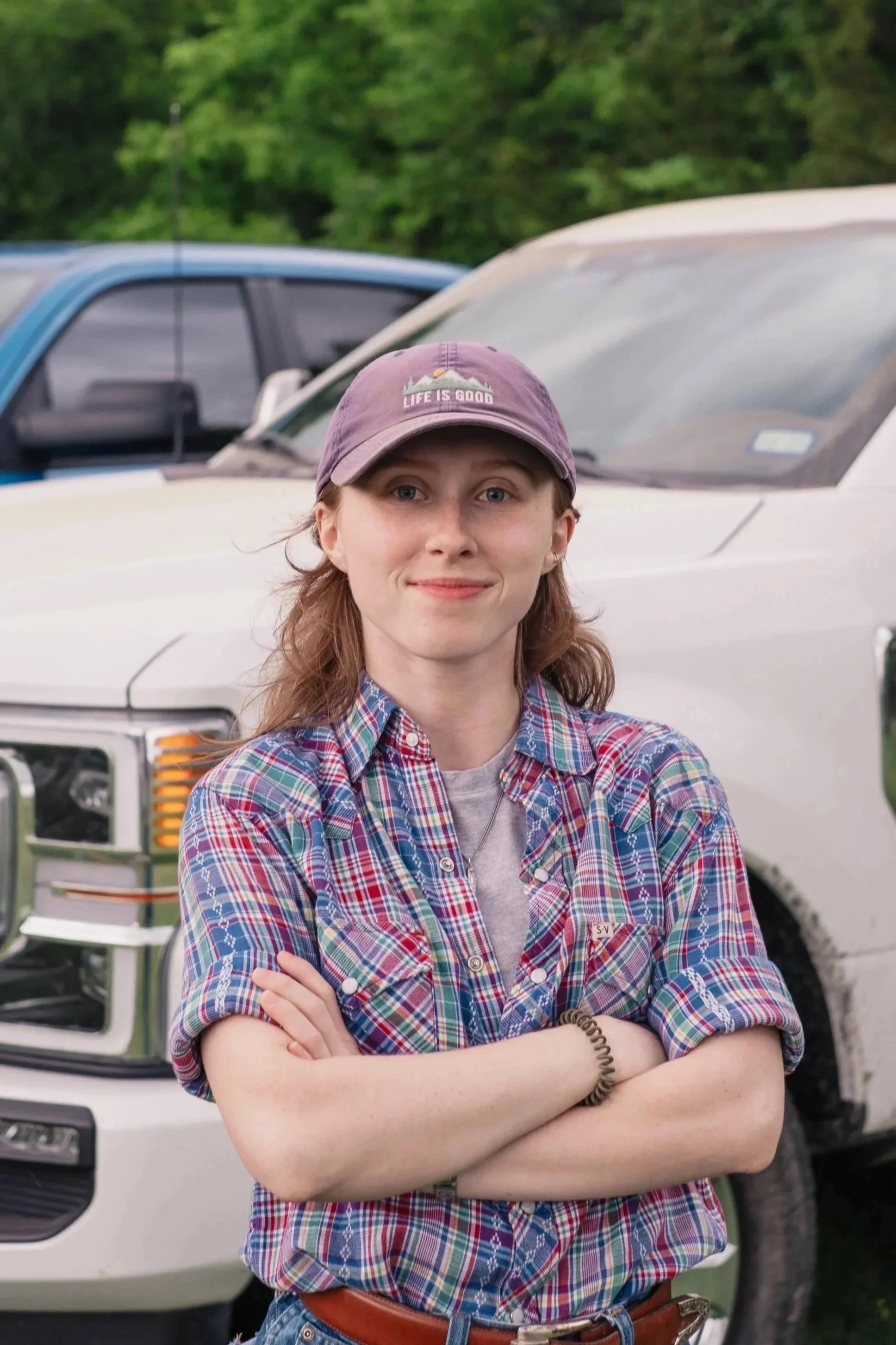 A young woman with light skin and brown hair wearing a purple baseball cap, a plaid shirt, and a brown belt, standing with arms crossed in front of a white vehicle, outdoors with green trees in the background.