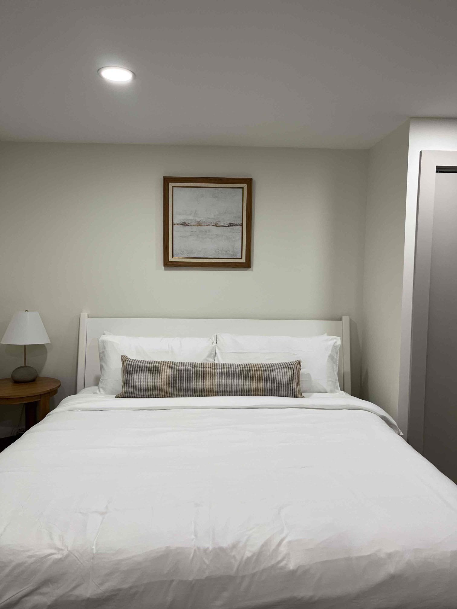 Finished basement bedroom with a clean white bed, wood nightstand, recessed lighting, and framed artwork above the headboard.