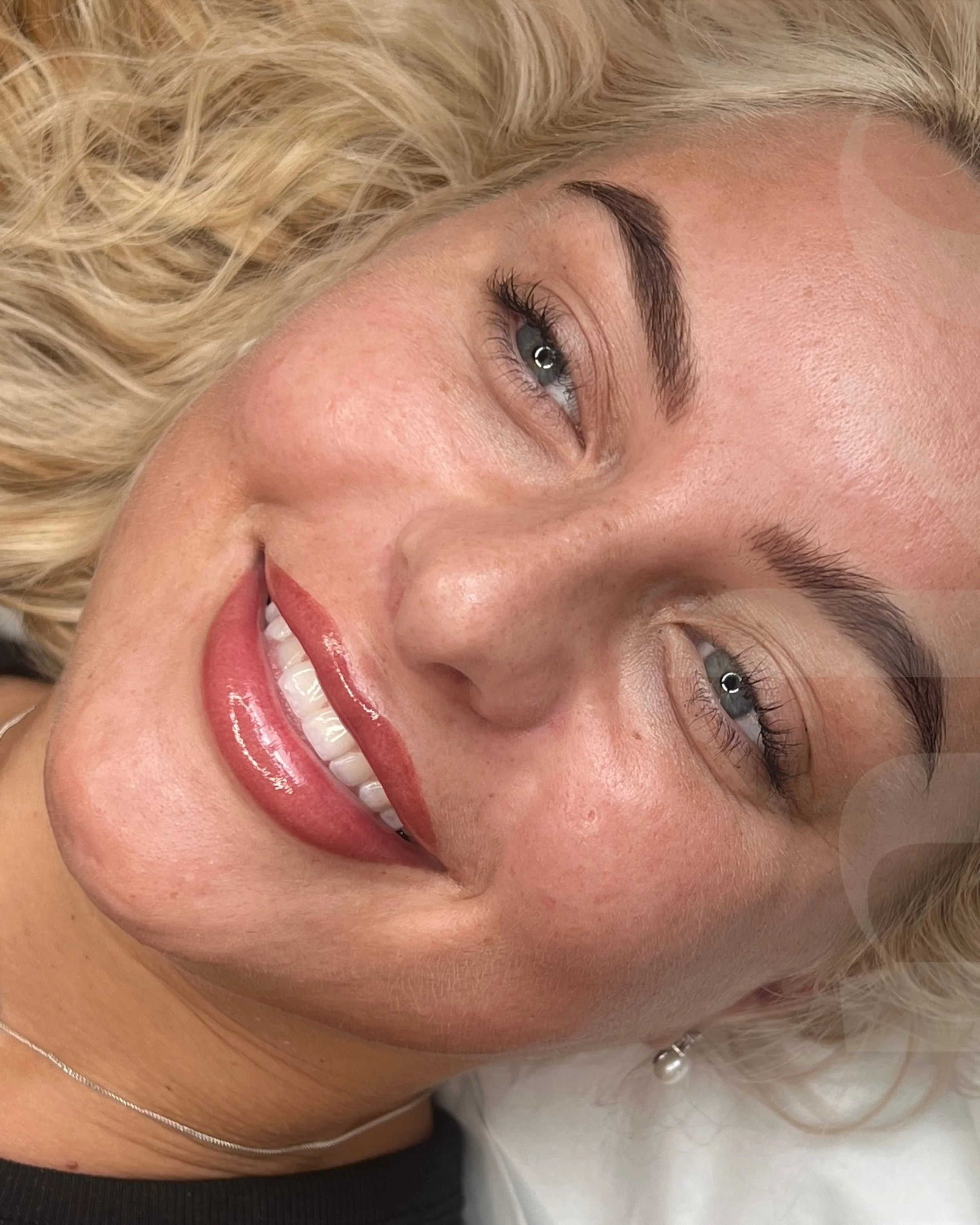 Close-up of a smiling woman with blonde curly hair, blue eyes, well-groomed eyebrows, and wearing makeup including lipstick and blush, lying down.