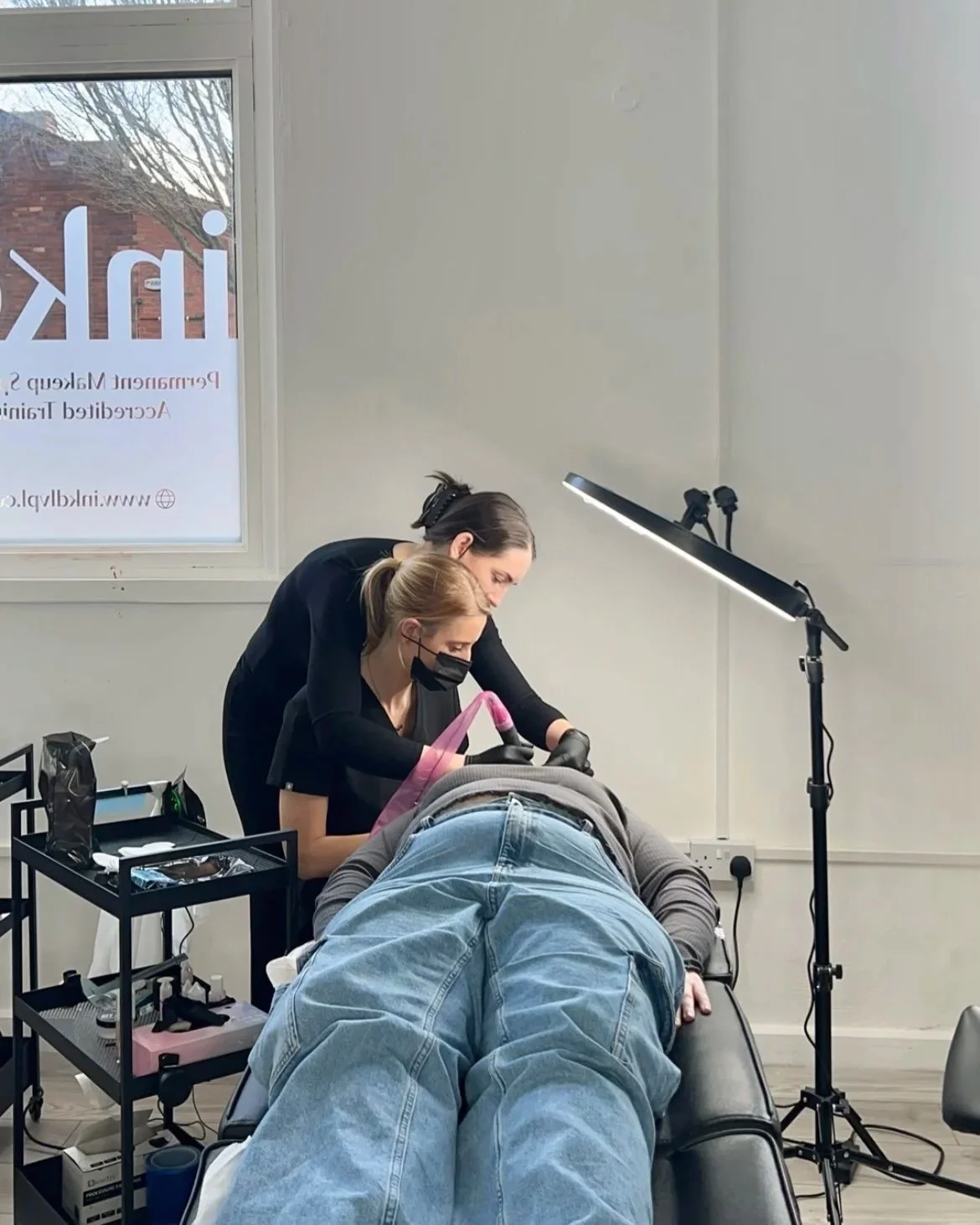 A tattoo artist is working on a client's arm in a well-lit room with a large window, two women are present, both wearing masks and gloves, with art supplies nearby.