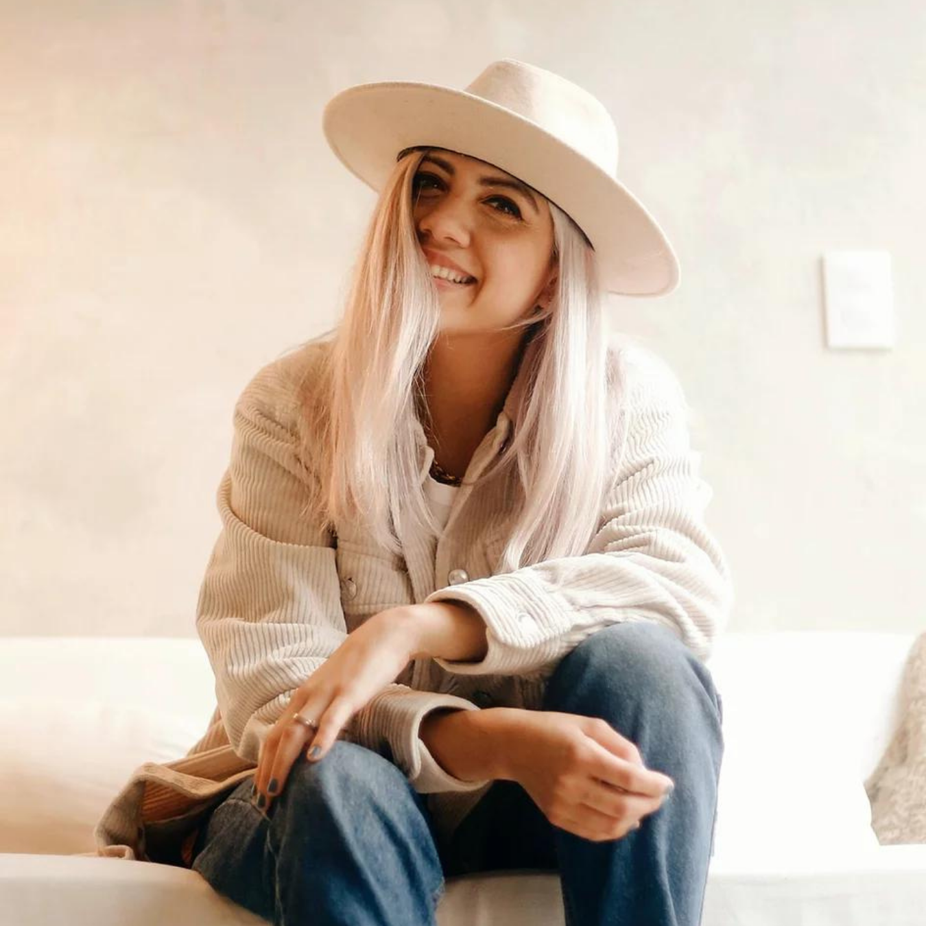 A young woman with long blonde hair, smiling, wearing a wide-brimmed beige hat, denim jeans, and a beige jacket, sitting on a white couch.