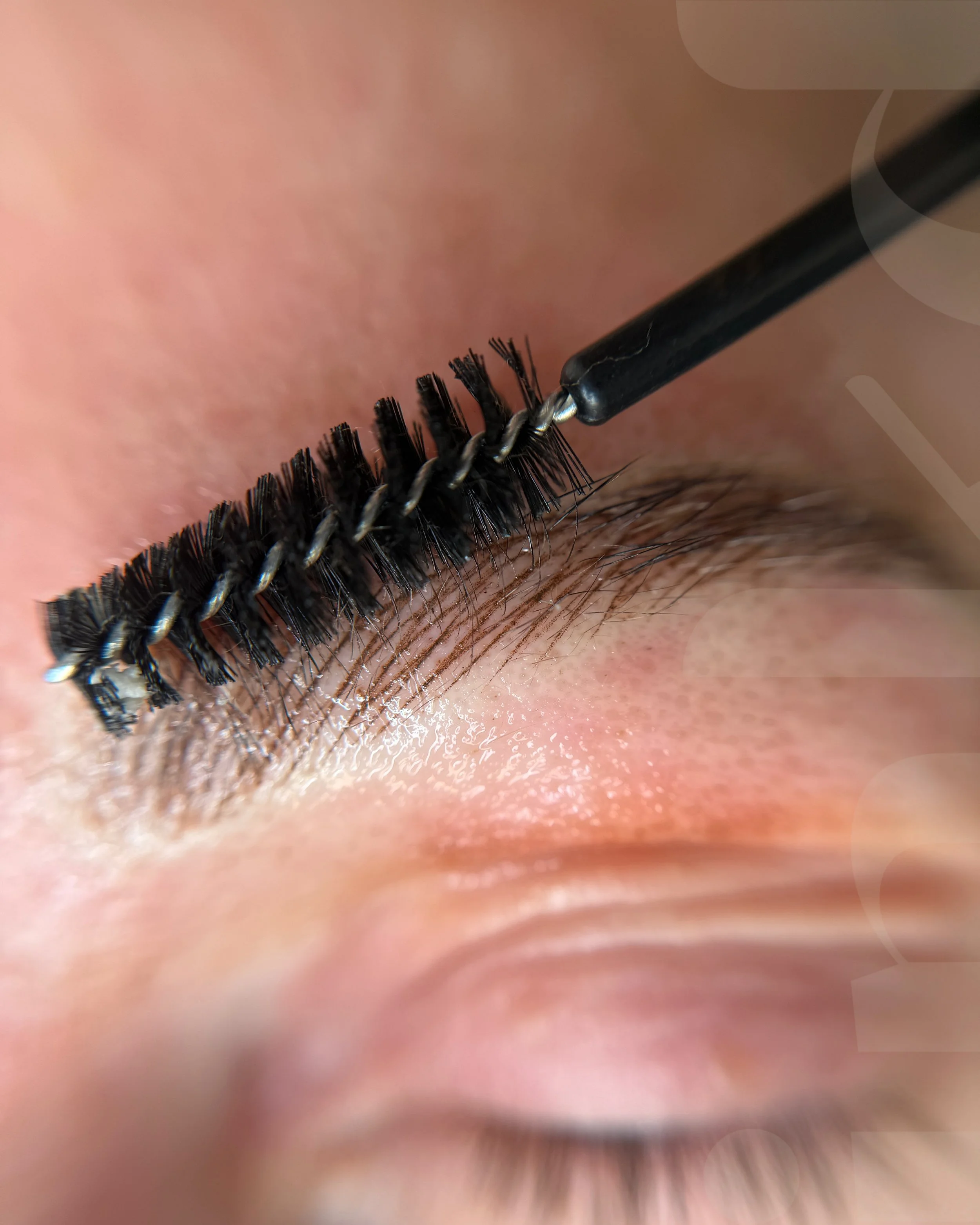 Close-up of a person's closed eye with eyelash extension application, showing a spoolie brush applying mascara or lash glue to the lashes.