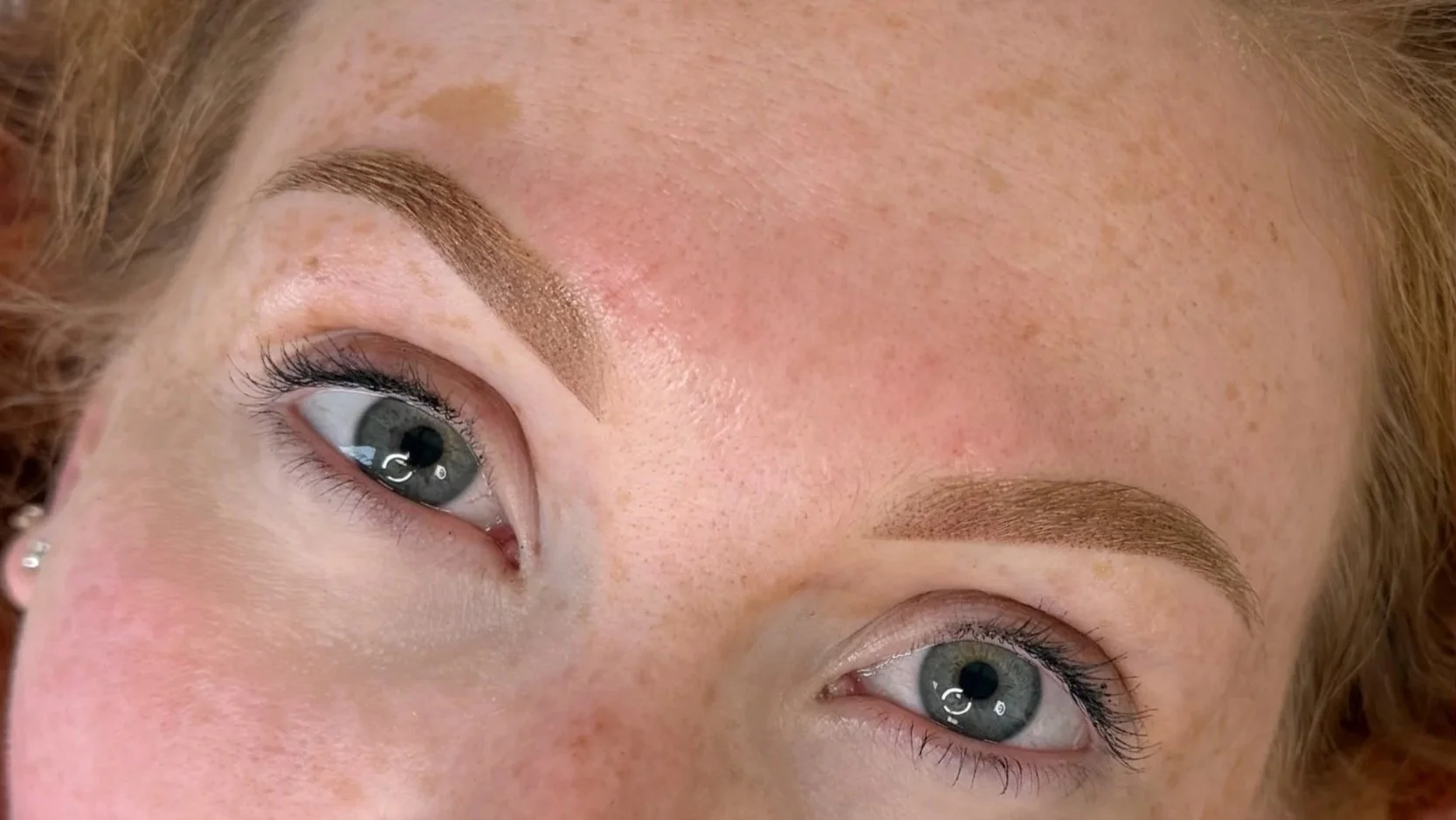 Close-up of a woman's face showing her eyes, eyebrows, and part of her forehead. The woman has light skin, blue eyes, and red hair. Her eyebrows have been shaped and filled in with makeup or microblading.