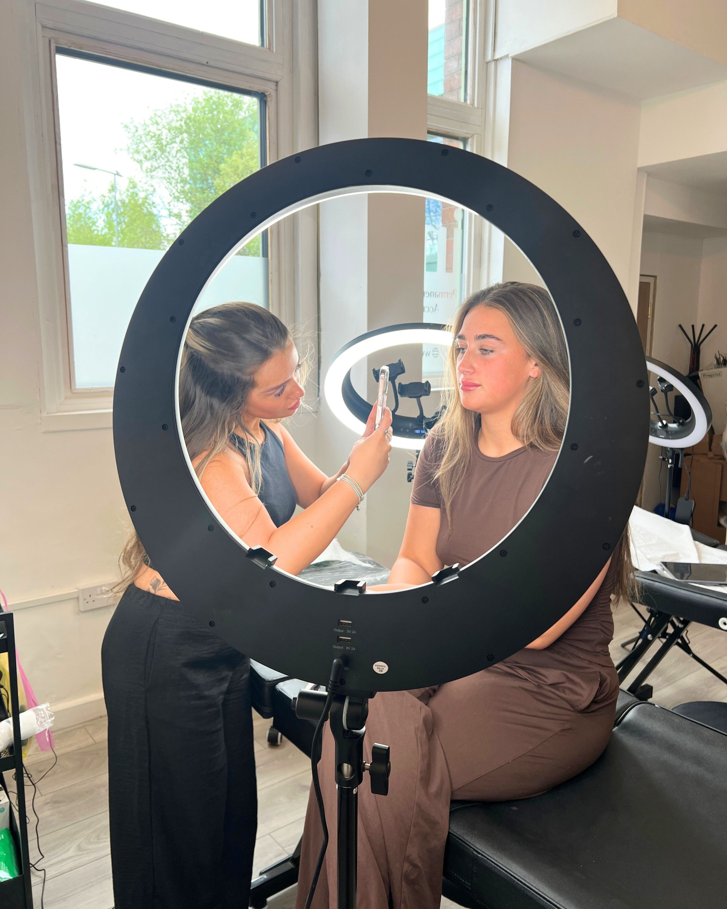 A makeup artist applies makeup to a woman seated on a black chair while taking a selfie with her phone inside a ring light. The room has large windows and additional ring lights in the background.