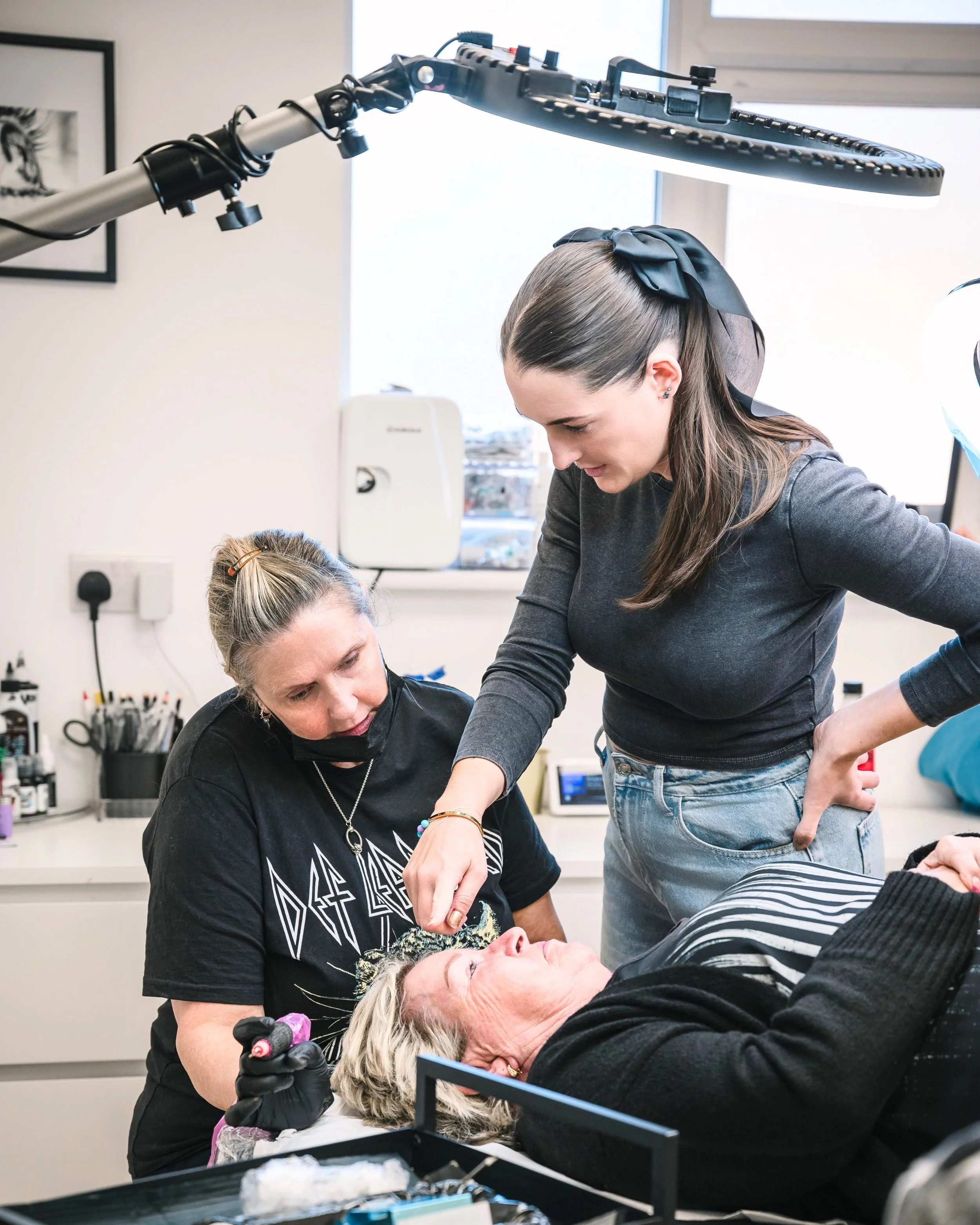 A woman lying on a clinic bed receiving a cosmetic or medical treatment from a technician while another woman watches closely in a clinical setting.