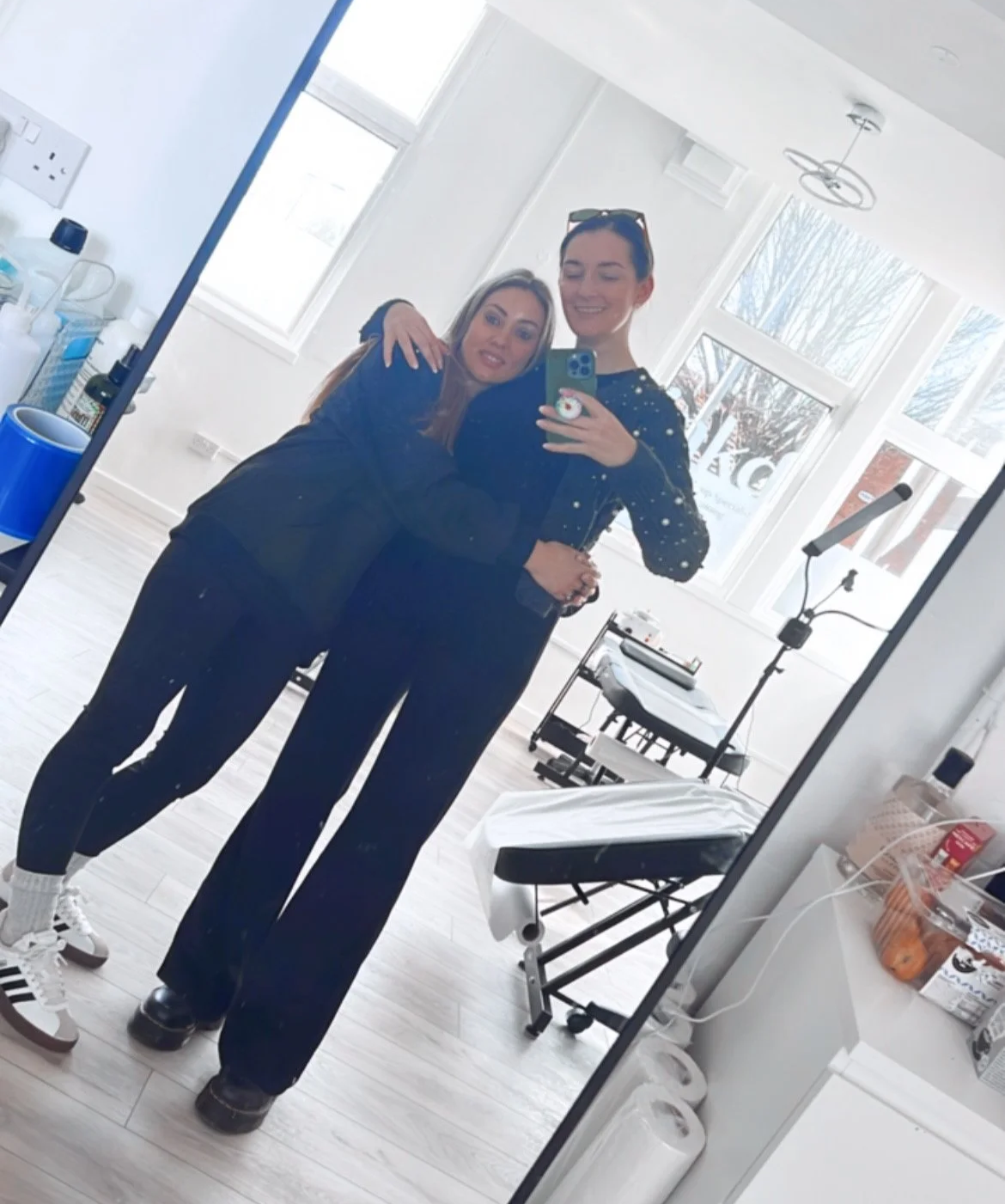 Two women taking a selfie in a bright room with large windows, medical equipment, and an examination table.