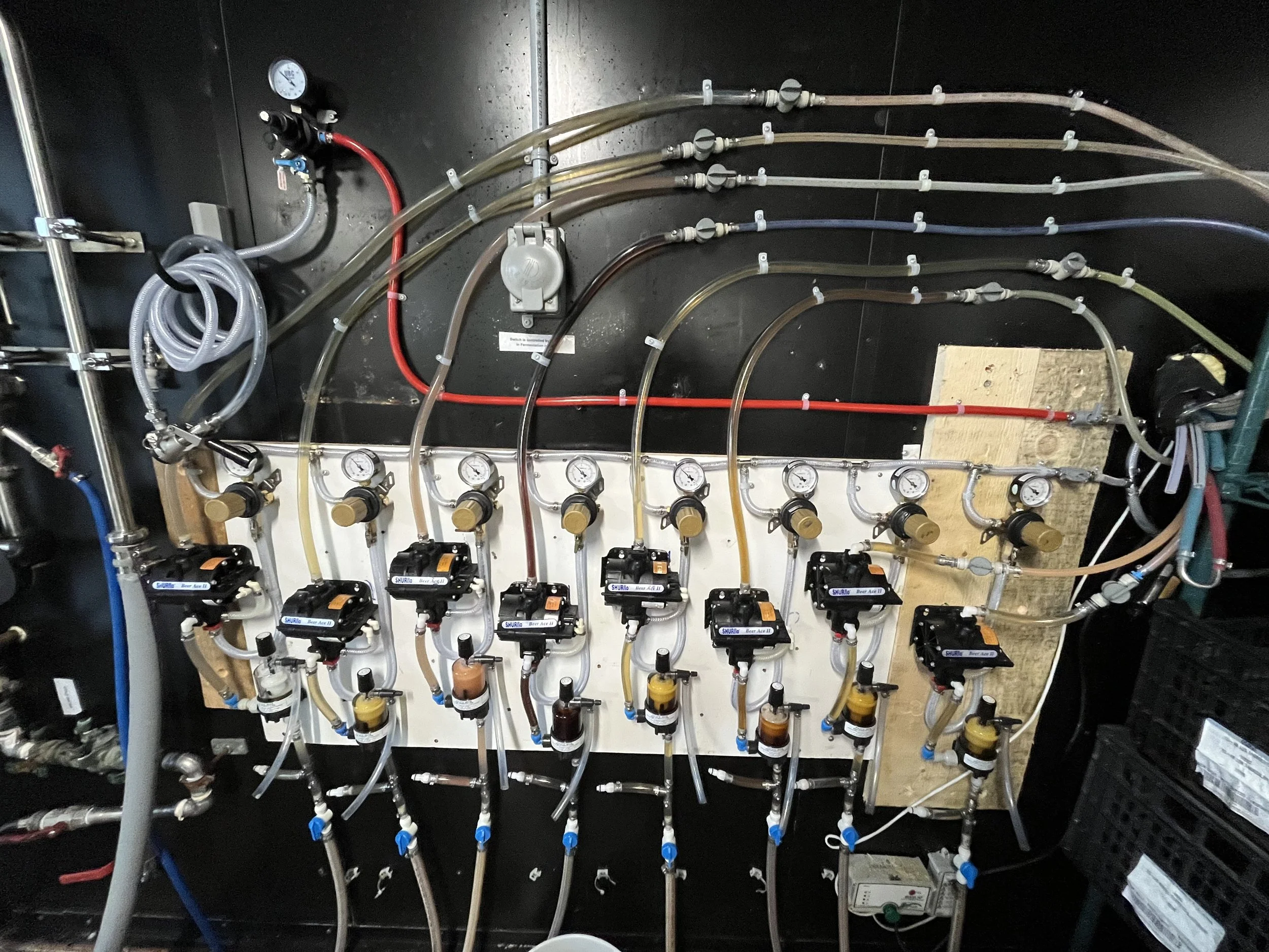 An intricate network of pipes, gauges, valves, and pumps mounted on a black wall, likely part of a scientific or industrial system.