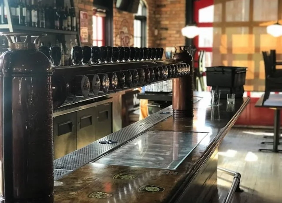 Empty bar counter with beer taps, glasses, and bar stools in a rustic pub or restaurant.