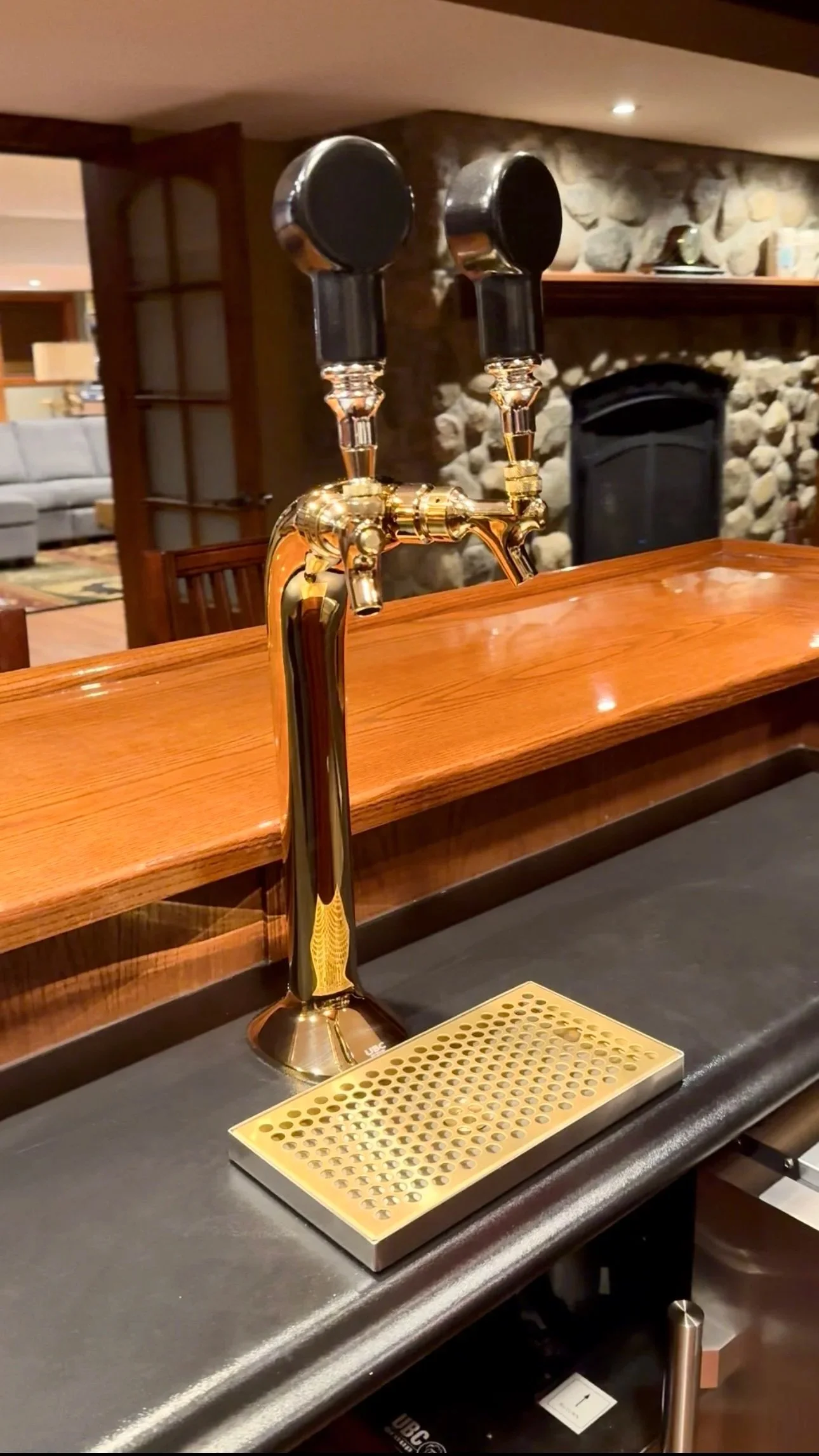 Close-up of a dual beer tap faucet with black handles and gold accents on a black countertop in a cozy home bar with a wooden counter and stone fireplace in the background.