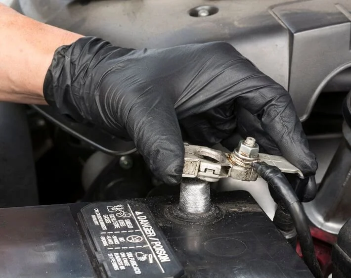 A person wearing black gloves handling a car battery cable connection.