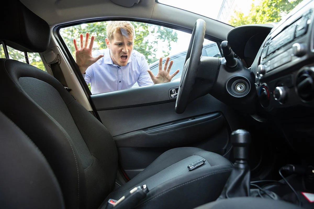 A man outside a car, looking frustrated or angry, pressing his hands against the window of the passenger side door.