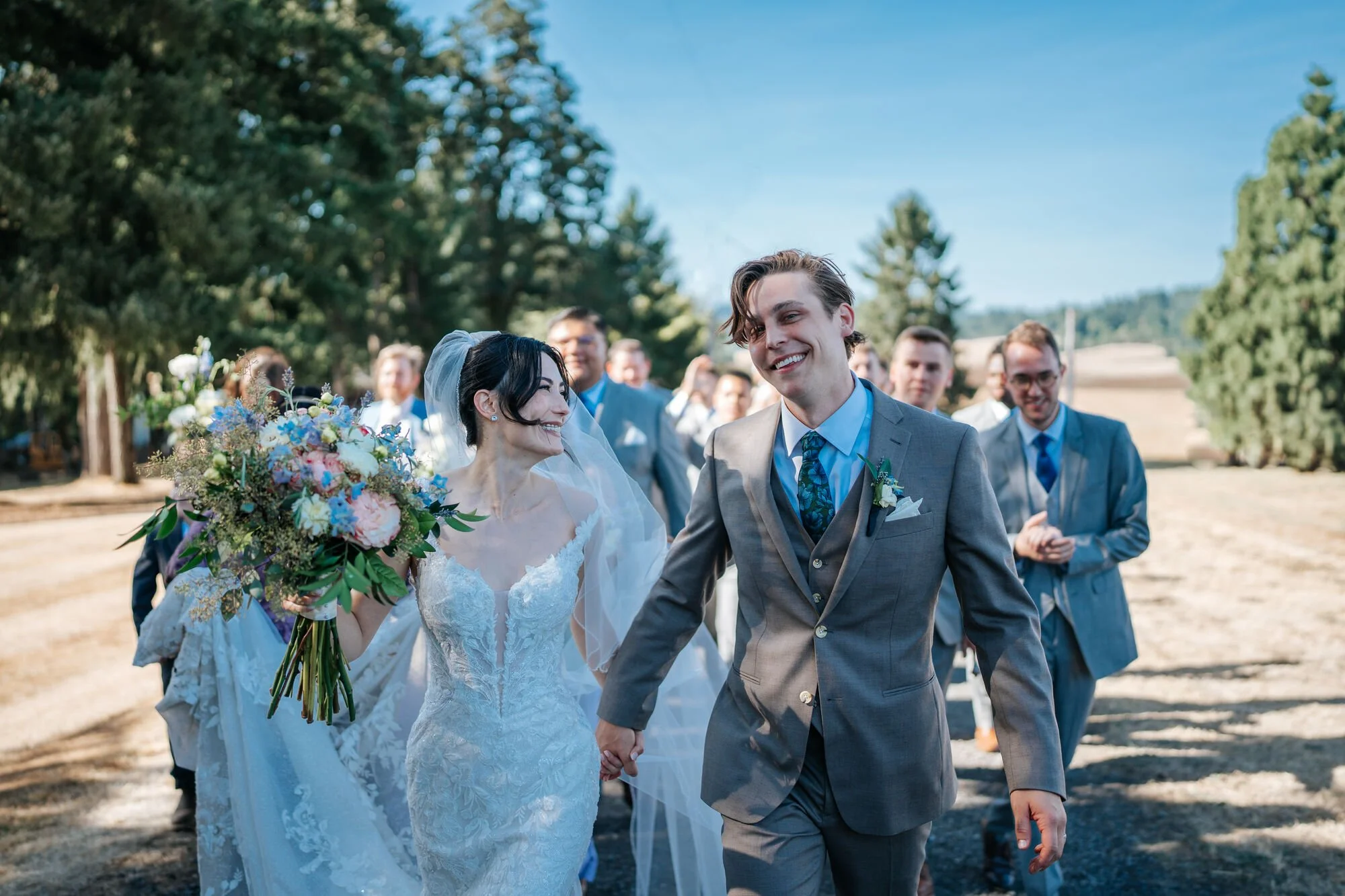 Bride and groom walk with their wedding party through a scenic Humboldt County setting. Fun candid group photo by Tex Kelly, McKinleyville and Fortuna wedding photographer.