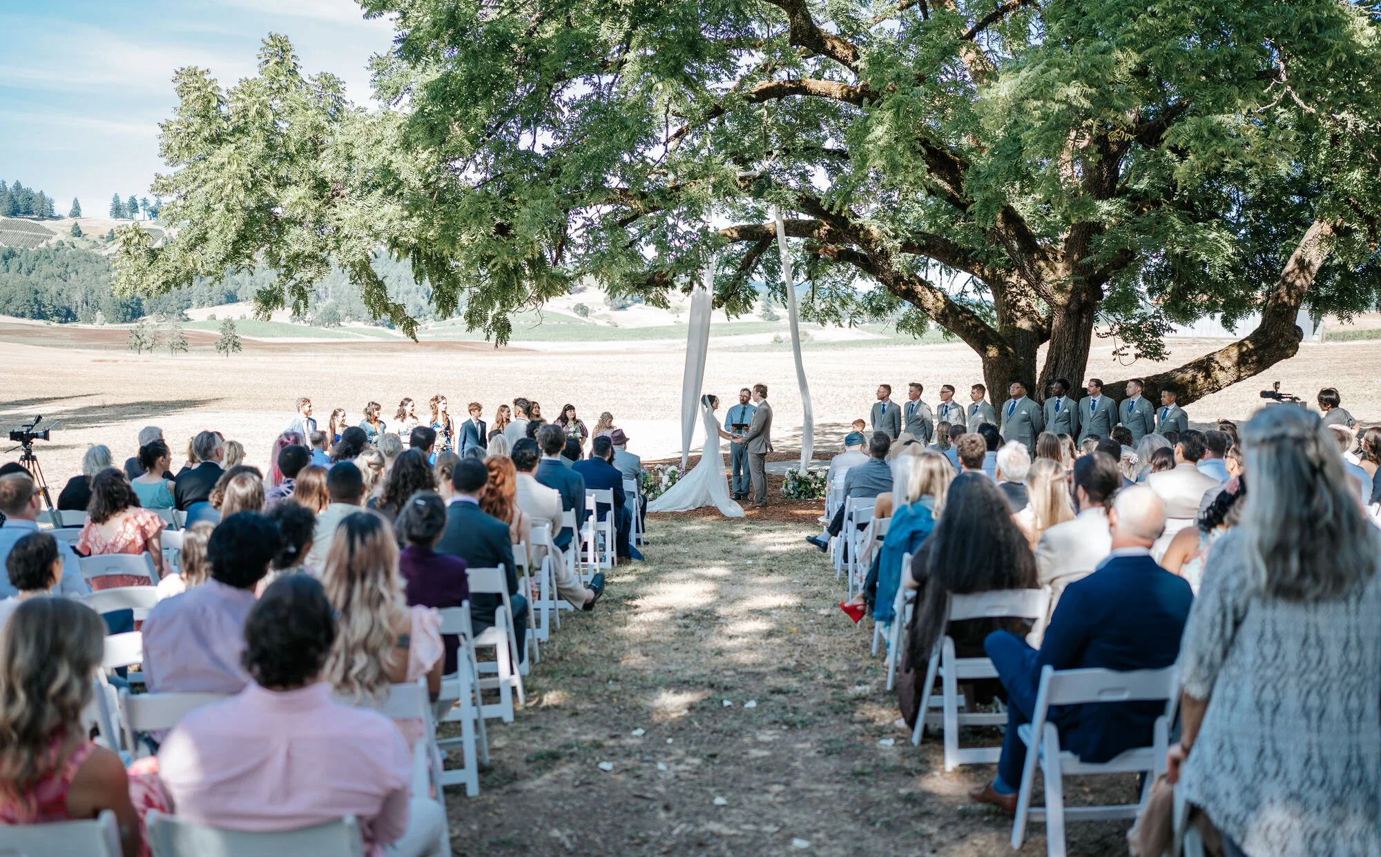 Tex Kelly Productions outdoor wedding ceremony photography in Humboldt County, California — guests seated under a large oak tree at a Fortuna wedding venue