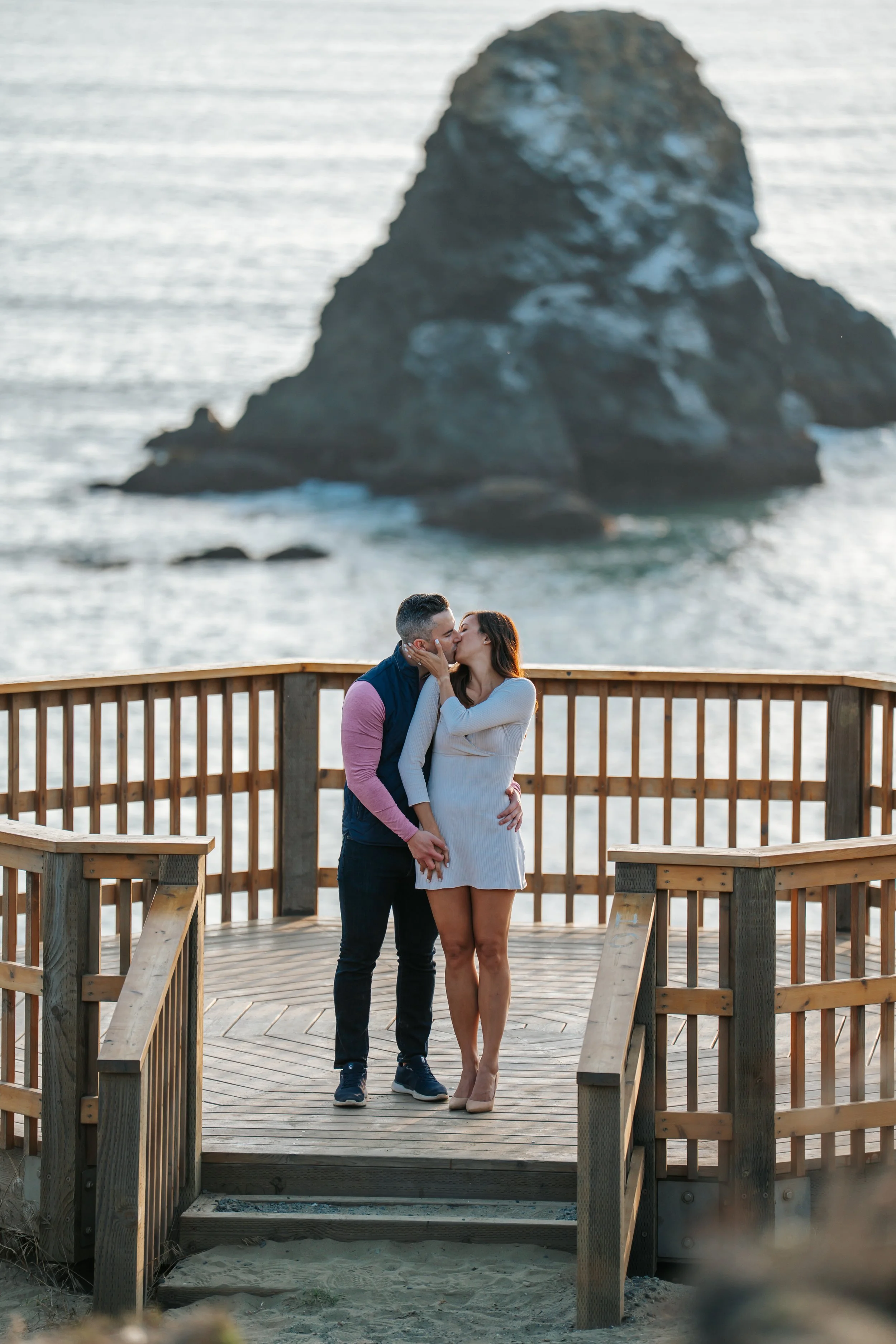 Engagement photography at Arcata oceanfront in Humboldt County California by Tex Kelly Productions - couple kisses on deck with dramatic sea stack and Pacific Ocean views