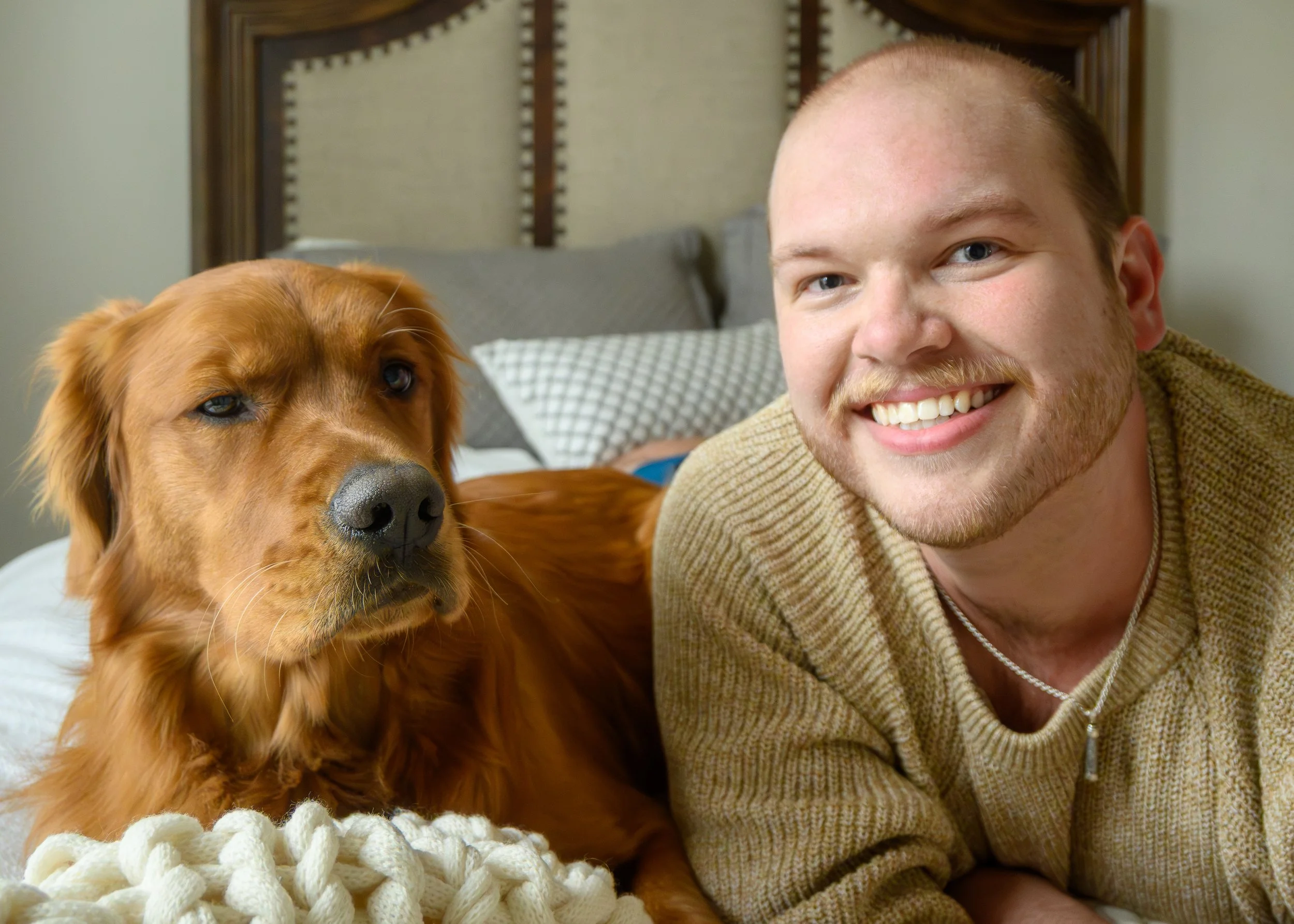 A smiling man with a beard and a dog lying on a bed, with pillows and a headboard in the background.