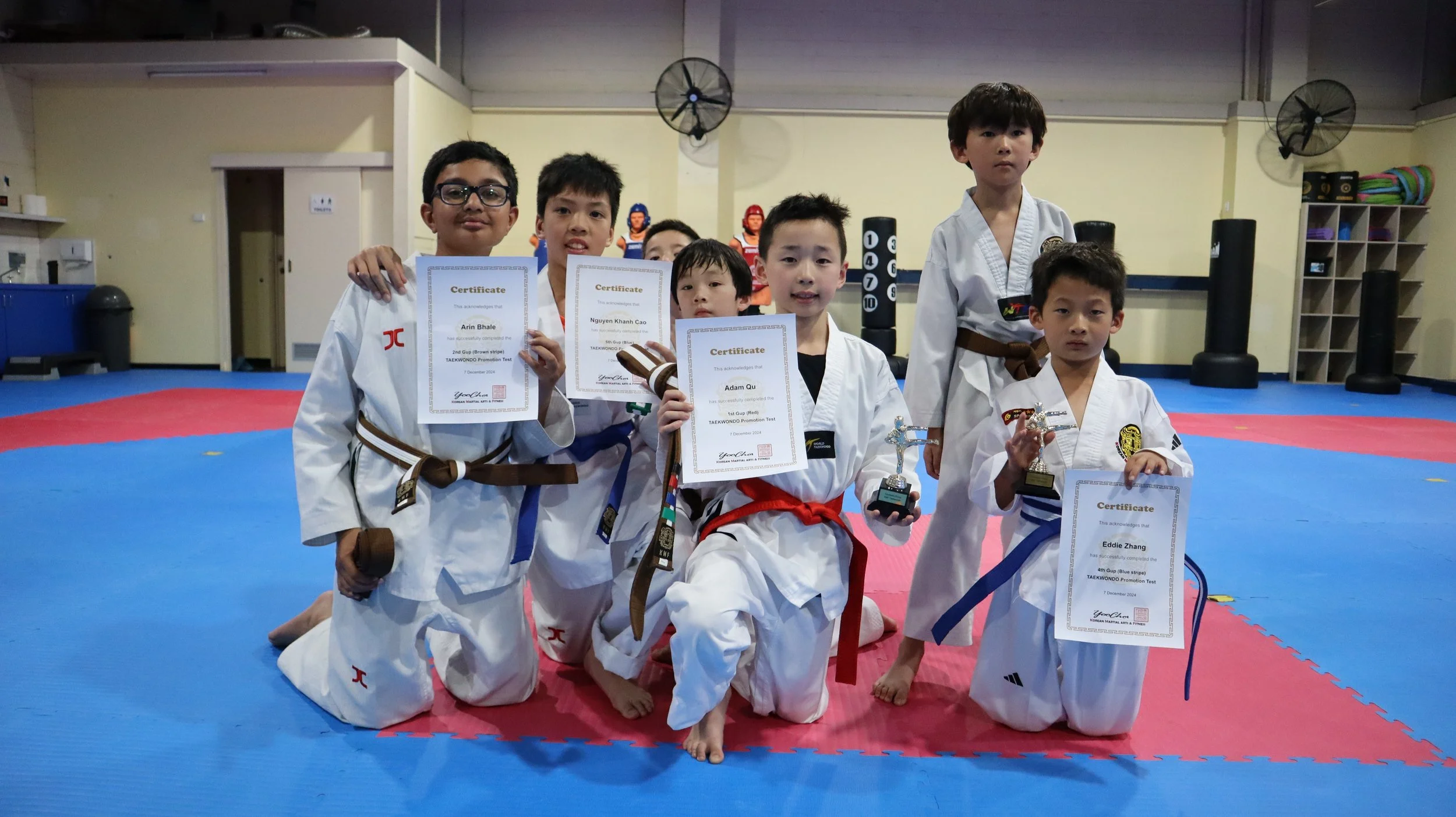 Group of young children dressed in white martial arts uniforms with colored belts, kneeling and standing on a martial arts training mat, holding certificates and trophies in a training facility.
