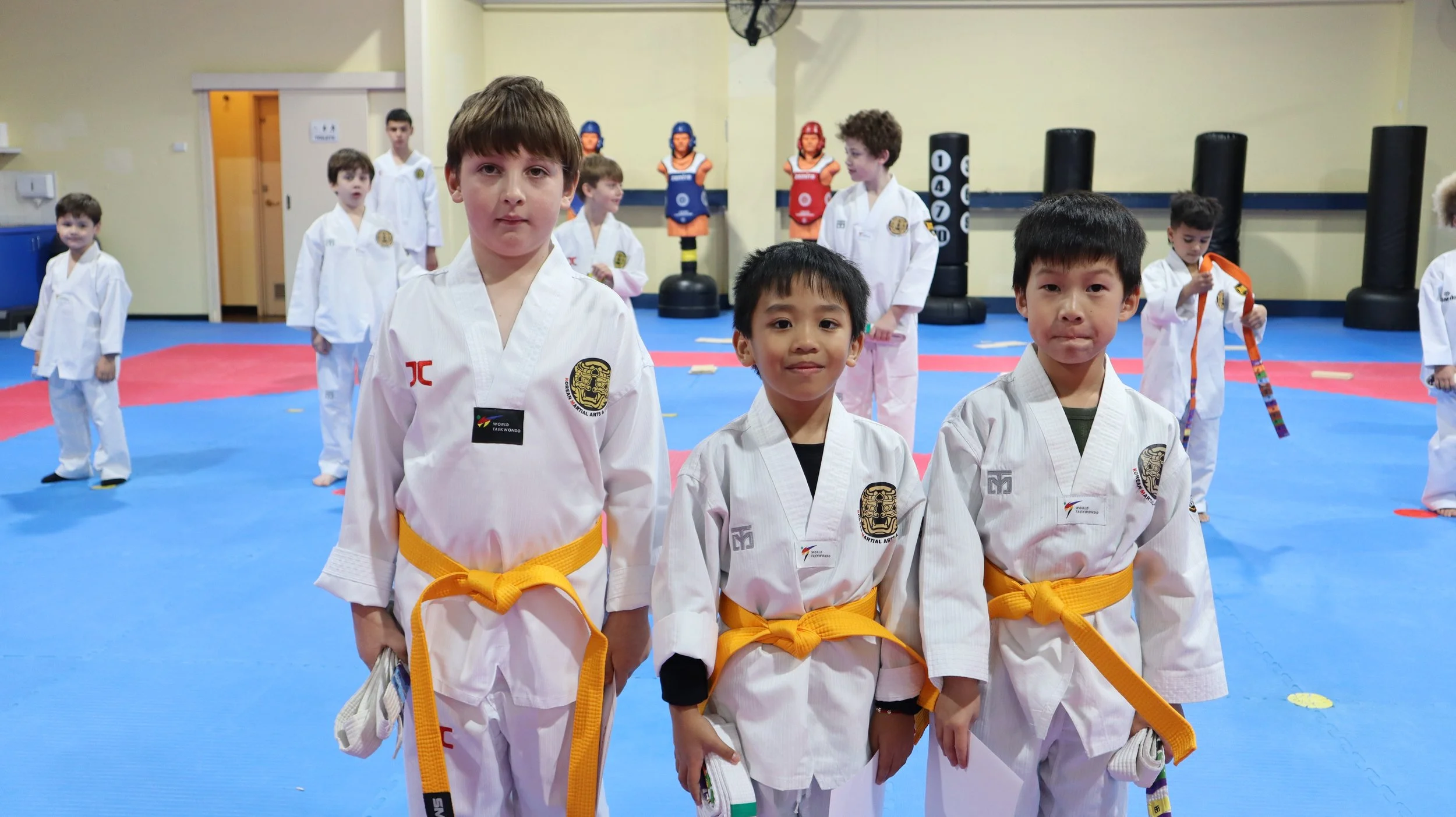 Kids in white martial arts uniforms with yellow belts standing in a dojo with other children and adults training in the background.