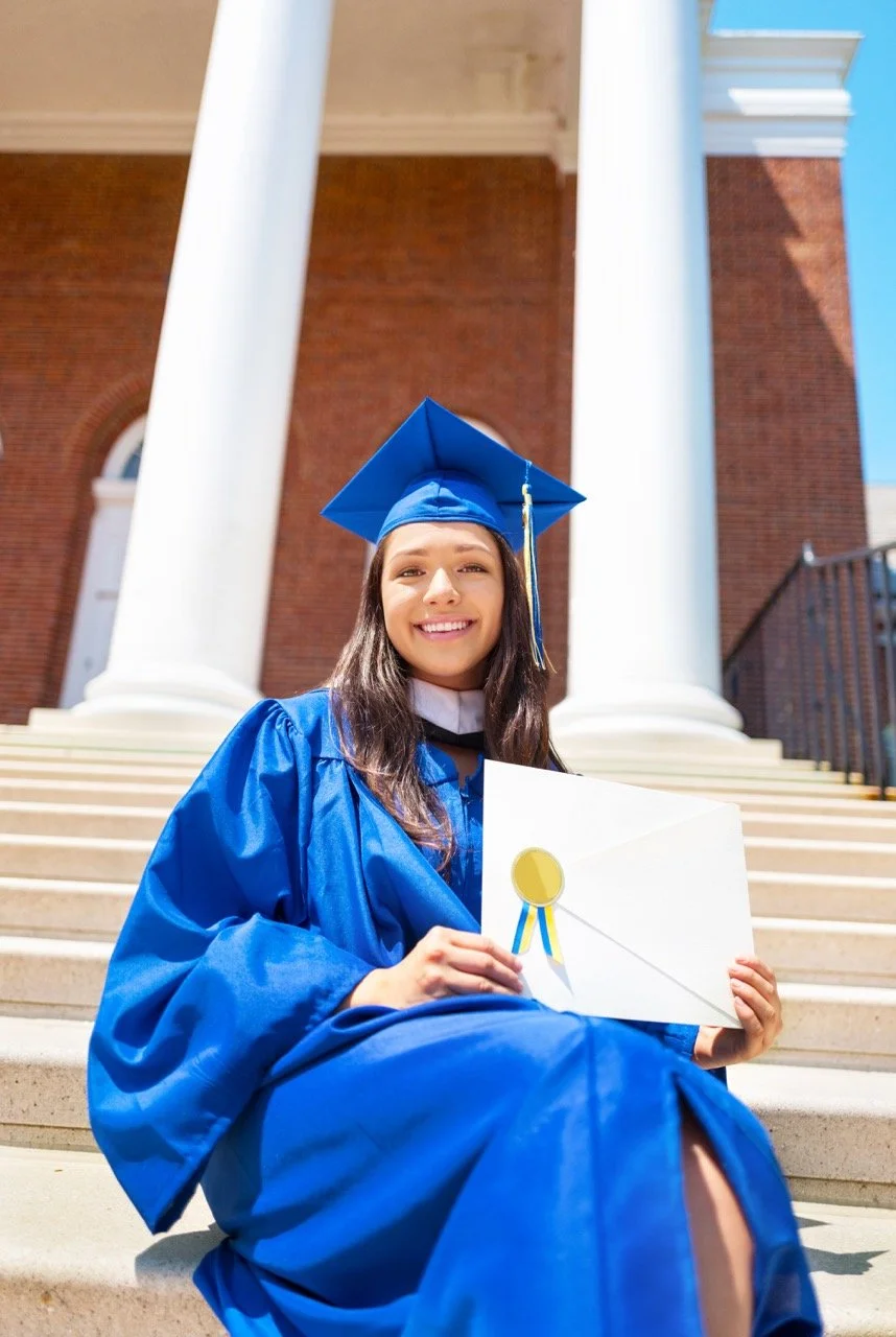A young woman in a blue graduation gown and cap sitting on outdoor steps holding an envelope with a gold medal sticker, smiling at the camera.